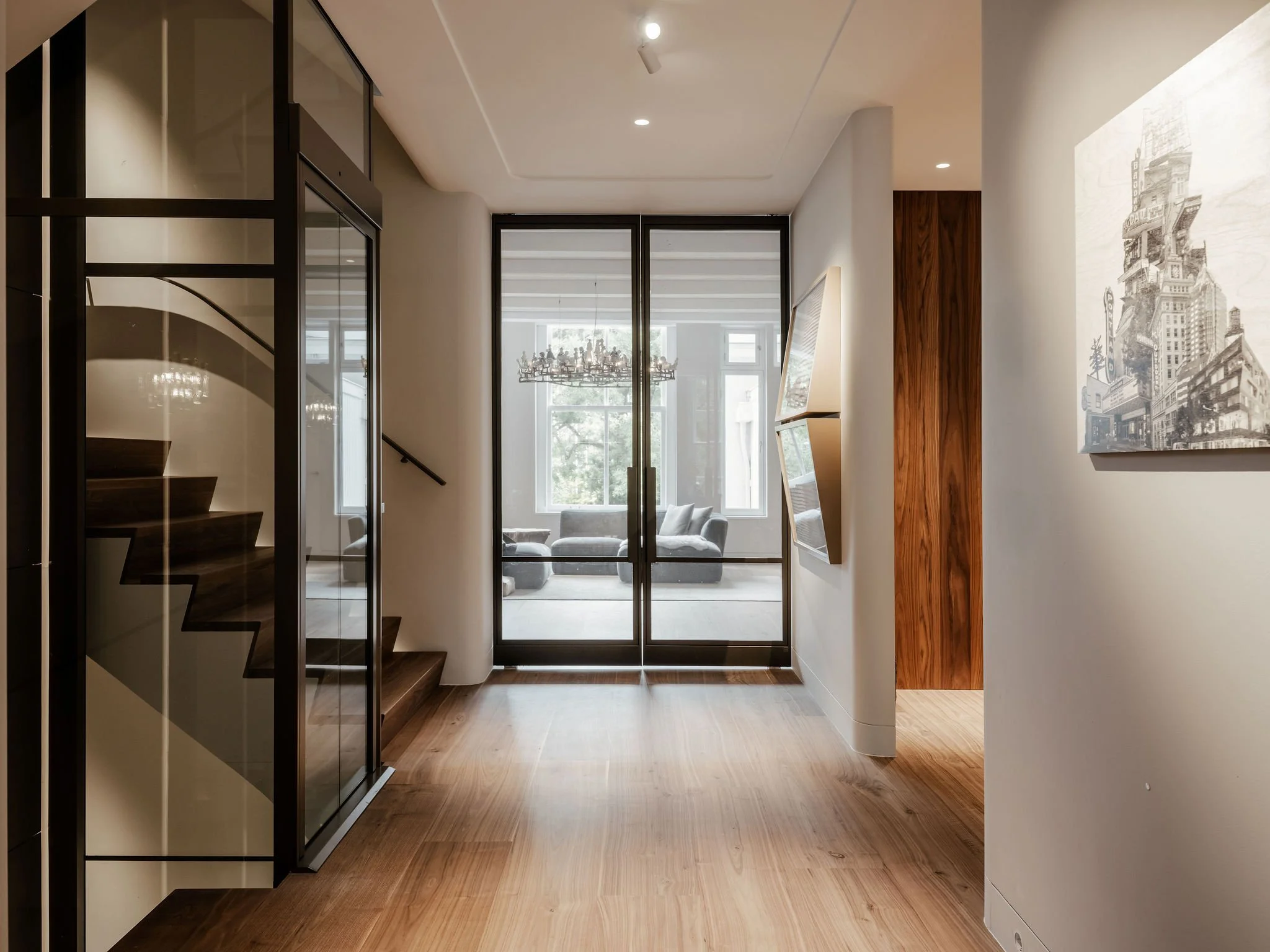 Modern hal met glazen deuren, houten trap, parketvloer en zicht op een lichte woonkamer.