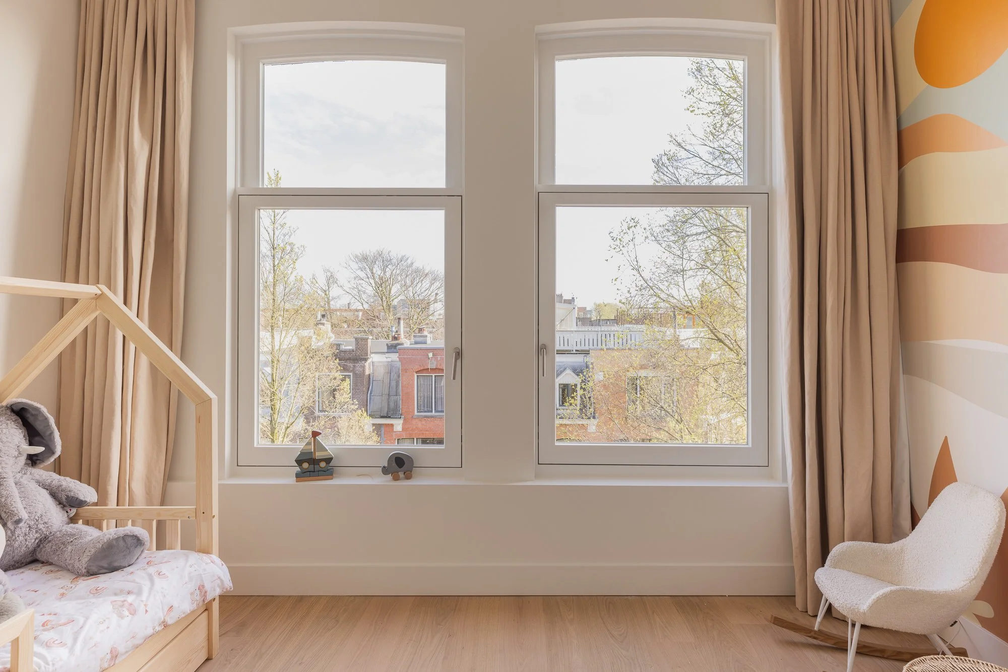 Lichte kinderkamer met groot raam, beige gordijnen, houten bed, knuffelbeer en witte schommelstoel.