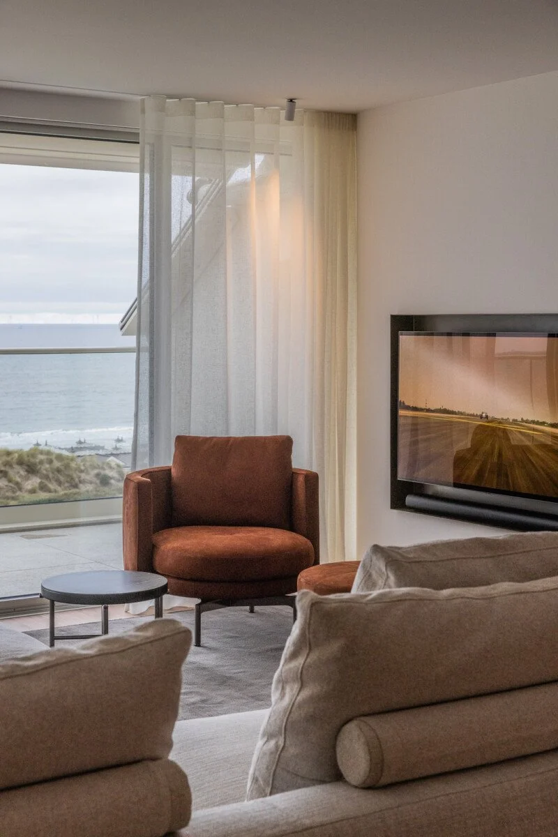 Moderne woonkamer met bruine fauteuils, beige banken, een salontafel en uitzicht op zee.