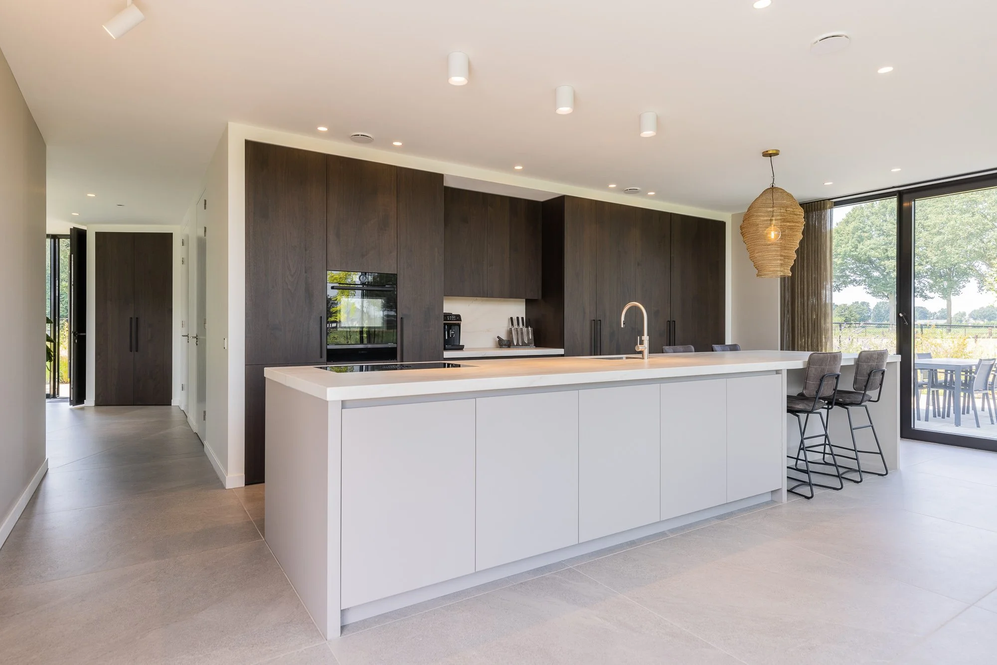 Moderne open keuken met groot wit kookeiland, donkere houten kasten, barstoelen en glazen deuren naar het terras