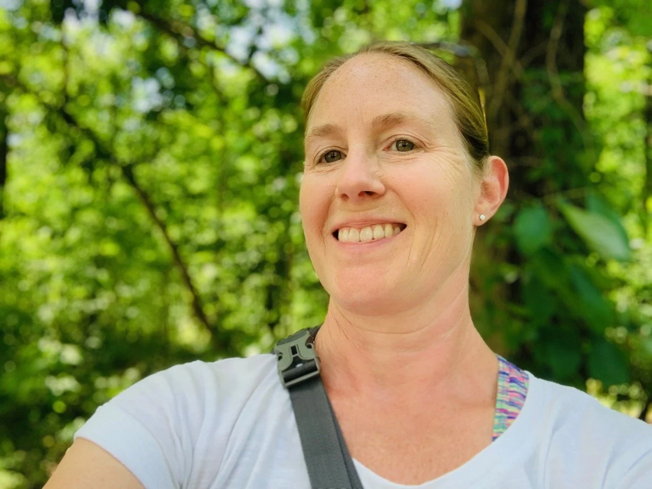 Sthira Sukham Yoga teacher Kristen Peterson wearing a white shirt with a grey strap over her shoulder, standing in front of lots of green trees and a blue sky.