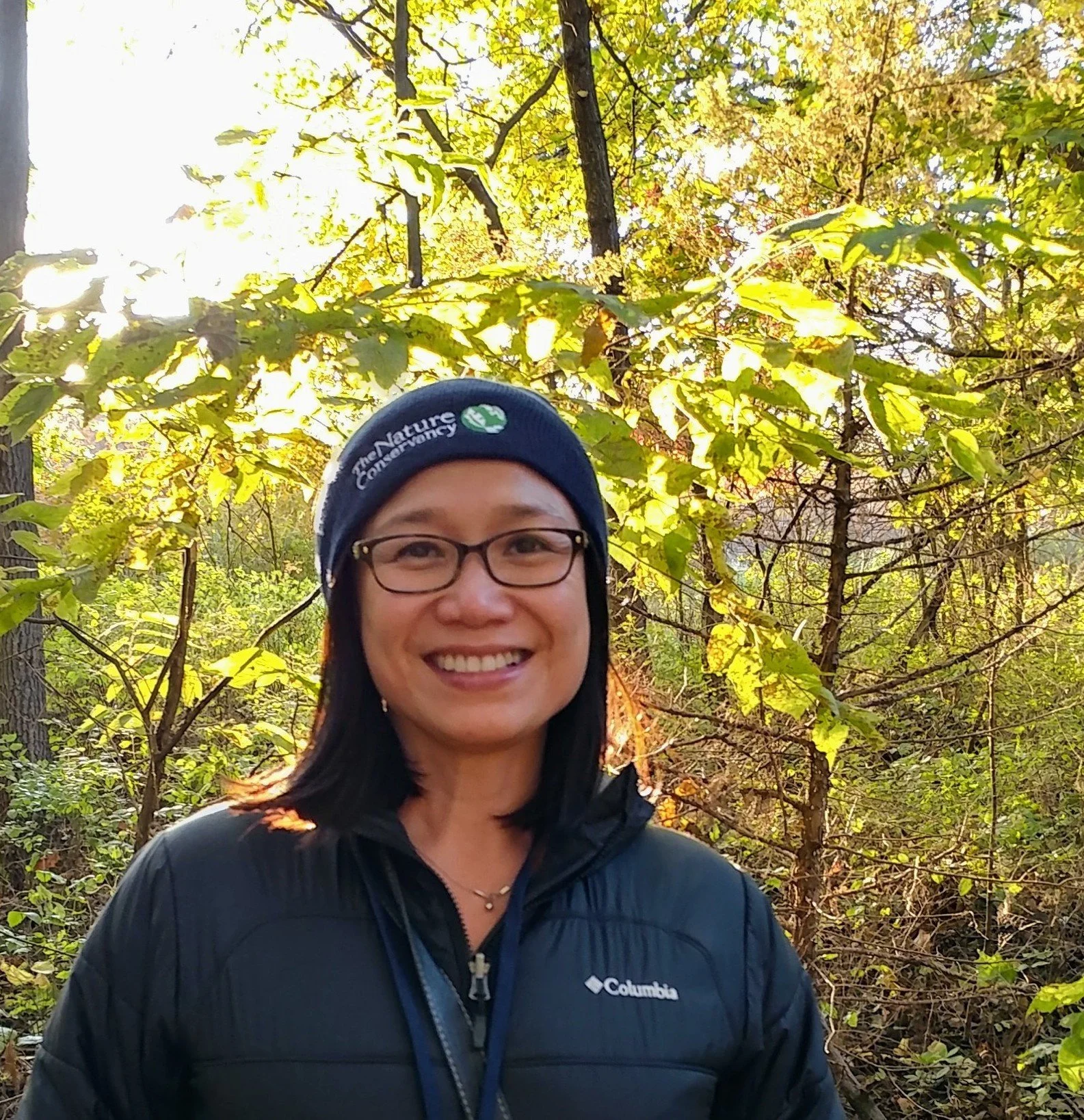 Sthira Sukham Yoga teacher Lucia Pham-Sinclair wearing dark framed glasses, a black jacket and a black beanie hat that reads the Nature Conservancy on it, surrounded by a lush green landscape.