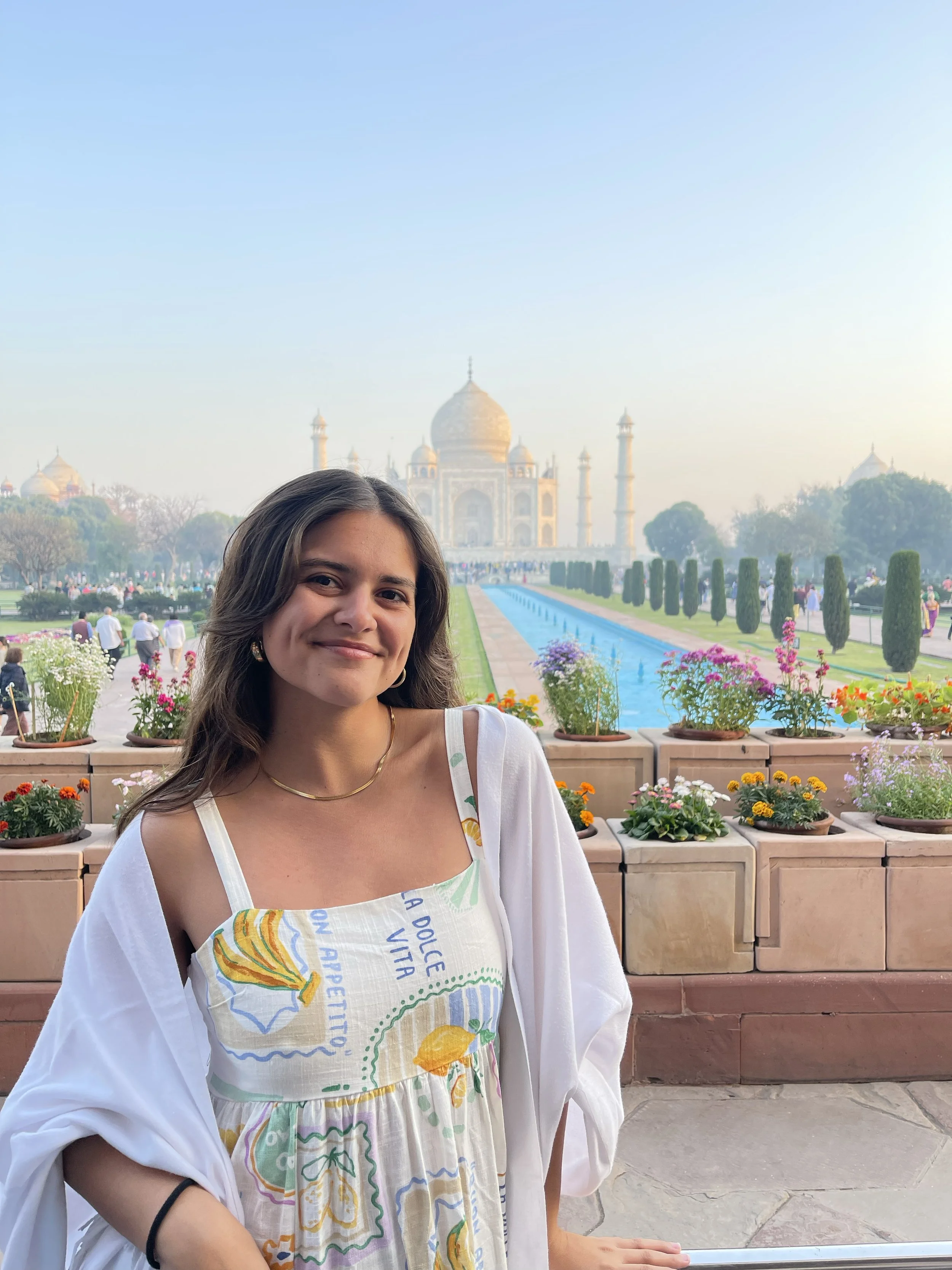 Sthira Sukham Yoga teacher Florencia Colon Perez stands in front of the Taj Mahal in India. She has long brown hair, a big smile and is wearing a patterned dress and white wrap over her shoulders. 