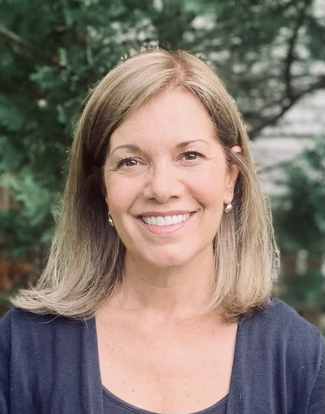 A picture of Sthira Sukham Yoga teacher Paula Hornberger, who has shoulder length light brown hair, beautiful brown eyes and a warm smile, and is wearing a navy blue sweater and tank top, standing in front of green trees. 