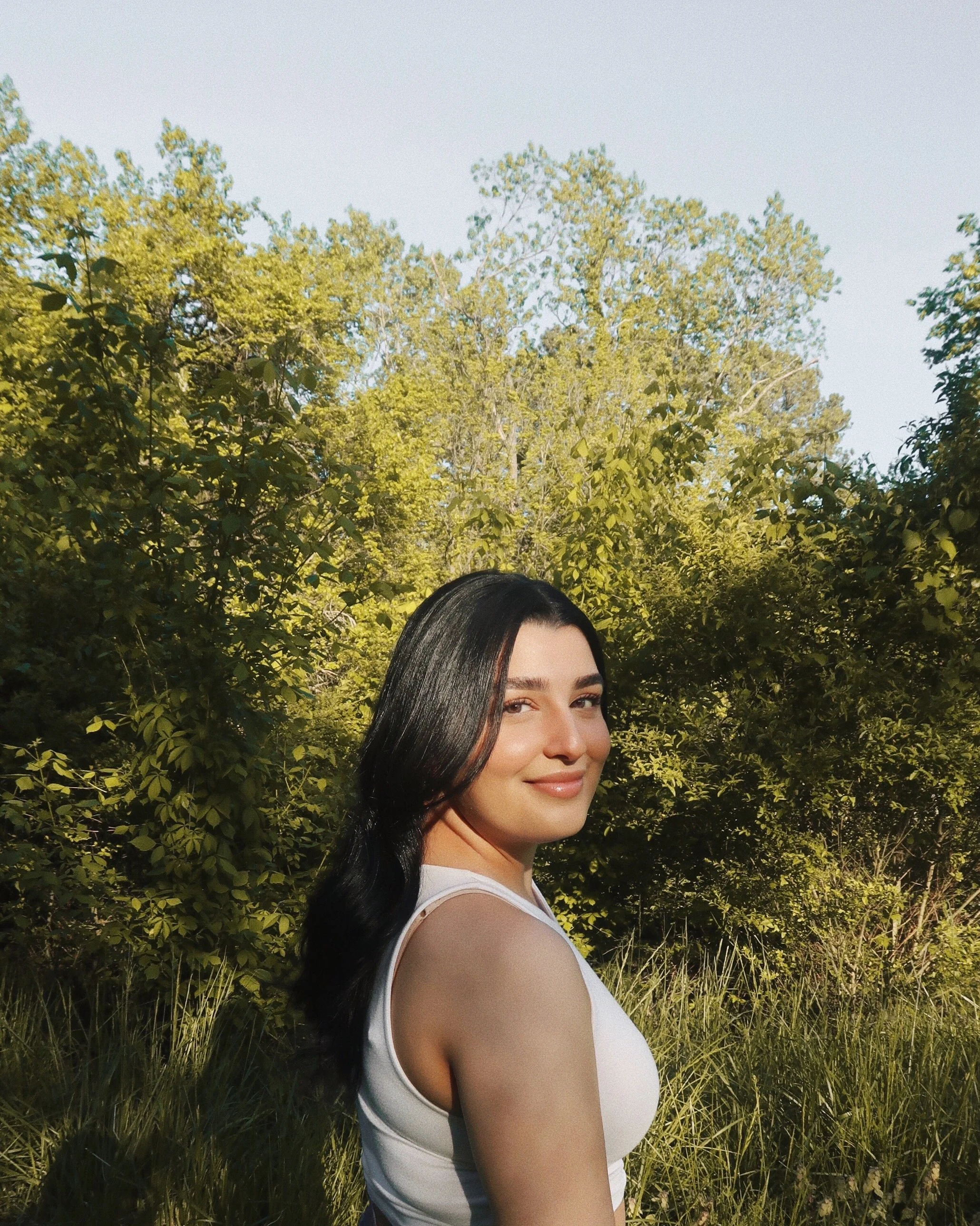 An image of Sthira Sukham Yoga, Restorative Yoga teacher Hadeel Al Weshahi who has long dark hair and is wearing a white tank top and is smiling warmly over her shoulder in a lush, sunlit forest.