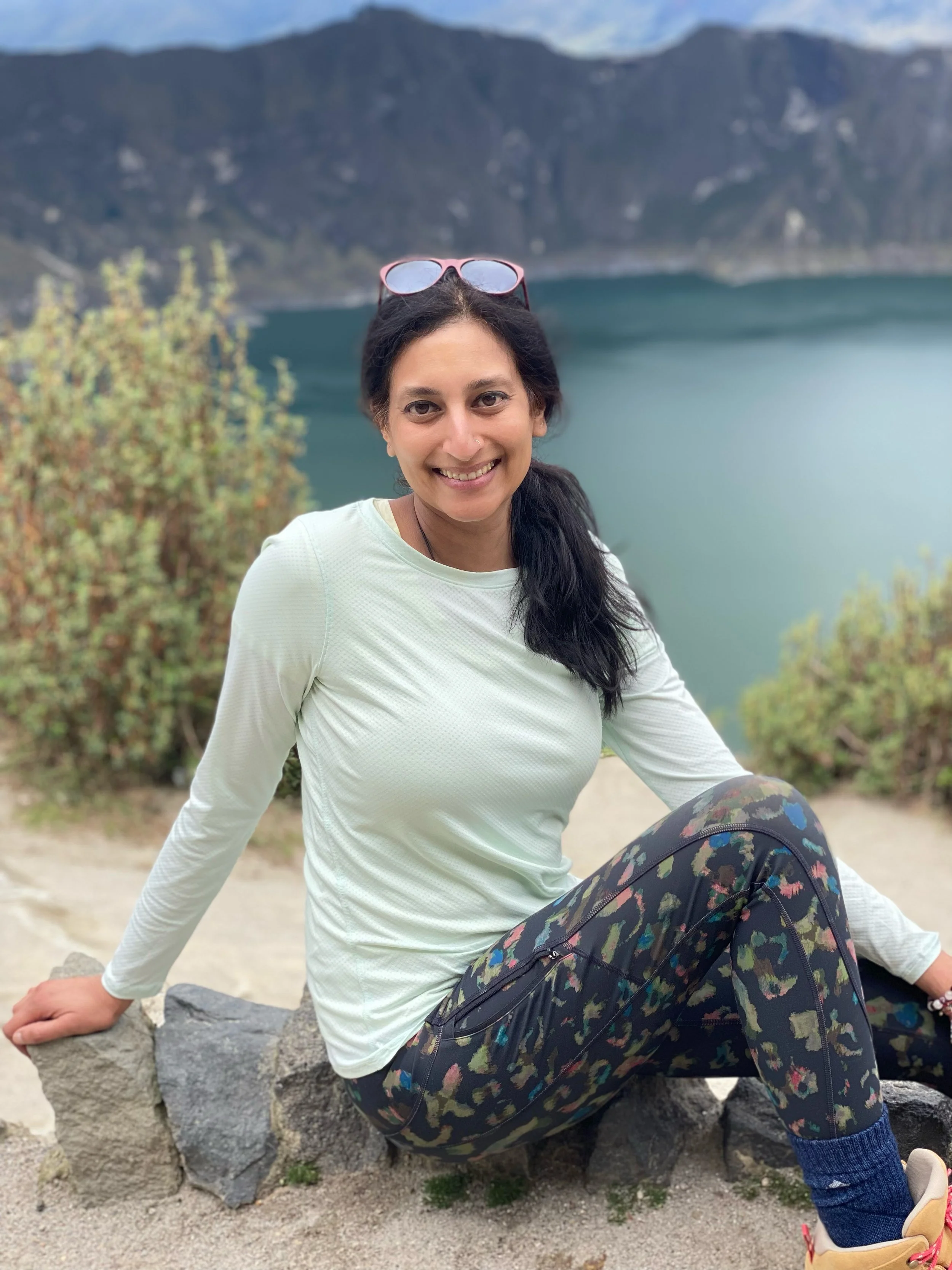 Sthira Sukham Yoga Hatha Flow teacher, Sarangi Bhatia sits in front of the Quilotoa Crater Lake, wearing a light green top and patterned leggings. She has long brown hair pulled back in a ponytail and has a warm, welcoming smile.