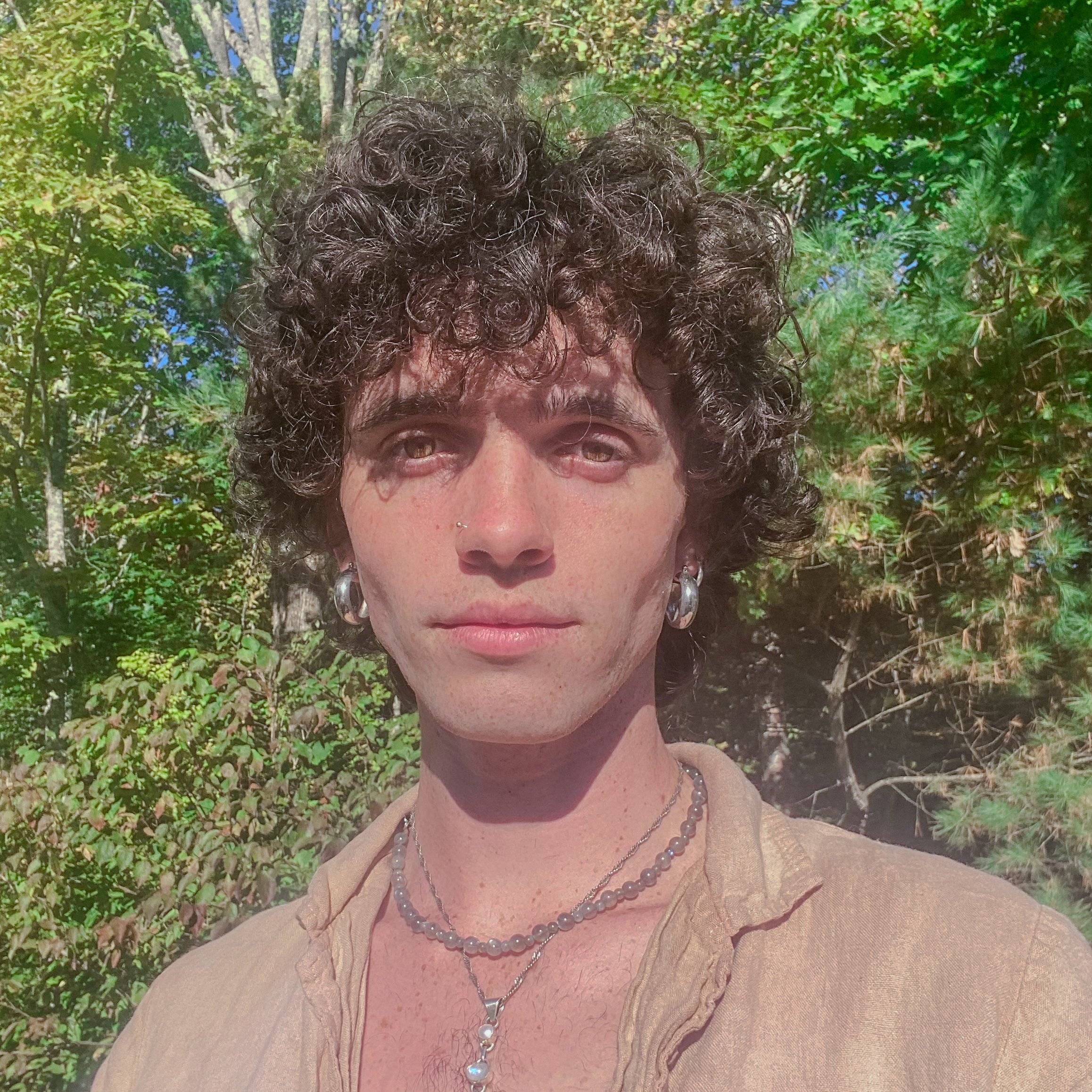 Sthira Sukham Yoga teacher Micah Doucette wears a tan colored shirt and a necklace. They have curly brown hair and are standing in front of a natural green background.