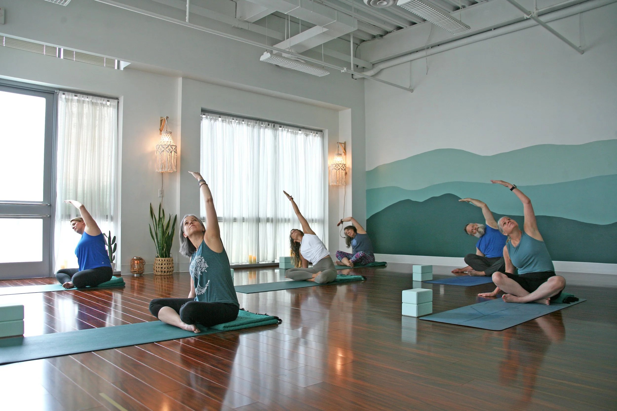 Adults enjoying gentle yoga at Sthira Sukham Yoga in Ashburn Virginia, practicing seated side stretches with optional yoga blocks in a quiet, supportive studio environment.