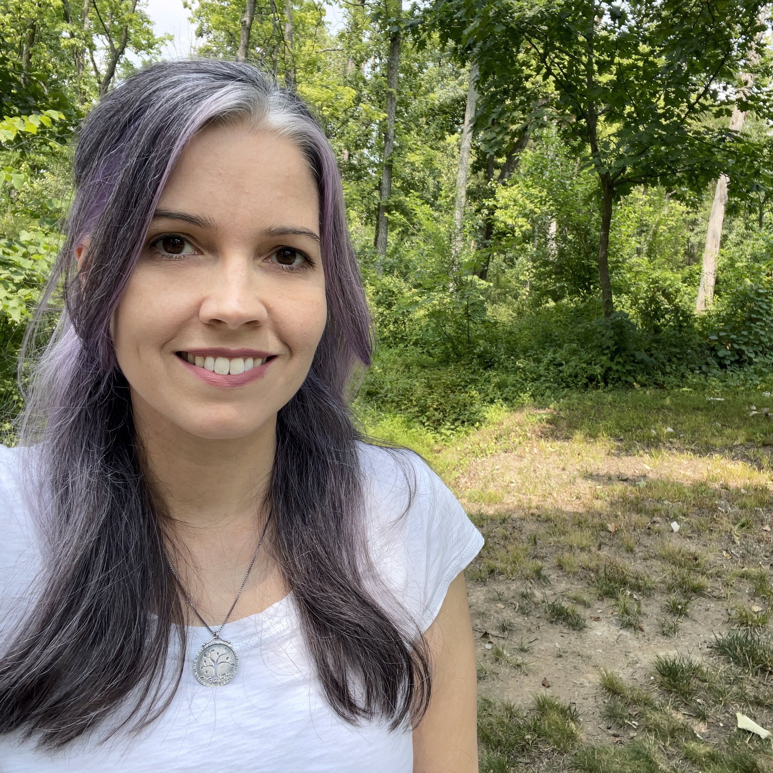 Sthira Sukham Yoga teacher Susan Quilty wearing a white t-shirt, and rocking light purple and silver hair stands in front of a backdrop of green trees and plants.