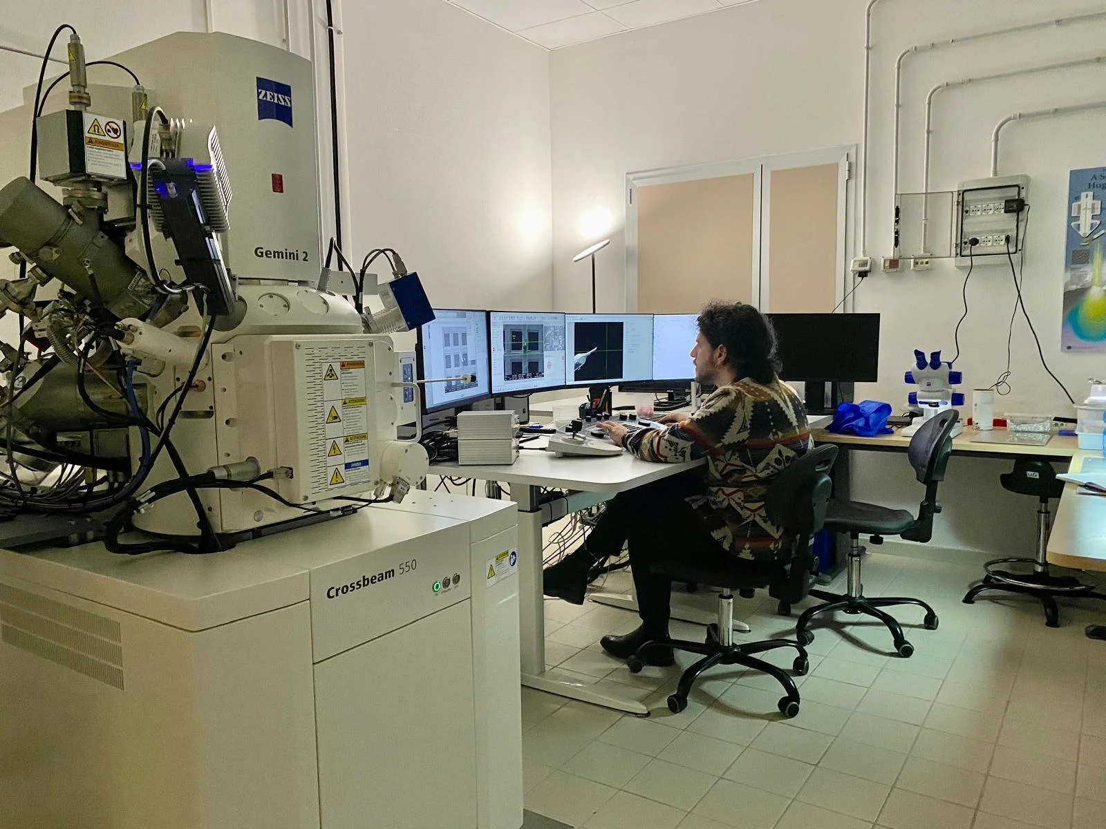 Luca sitting at a desk with five monitors in a lab with an electron microscope in the foreground. Images on the monitors show the micrographs being collected with the microscope.