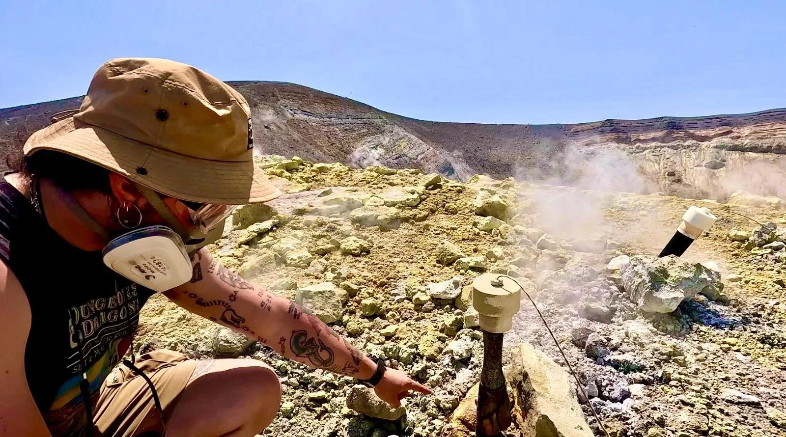 Luca wearing a ventilator sitting down near a sampling port in a volcanic fumarole. The rocks around are stained yellow from sulfur.