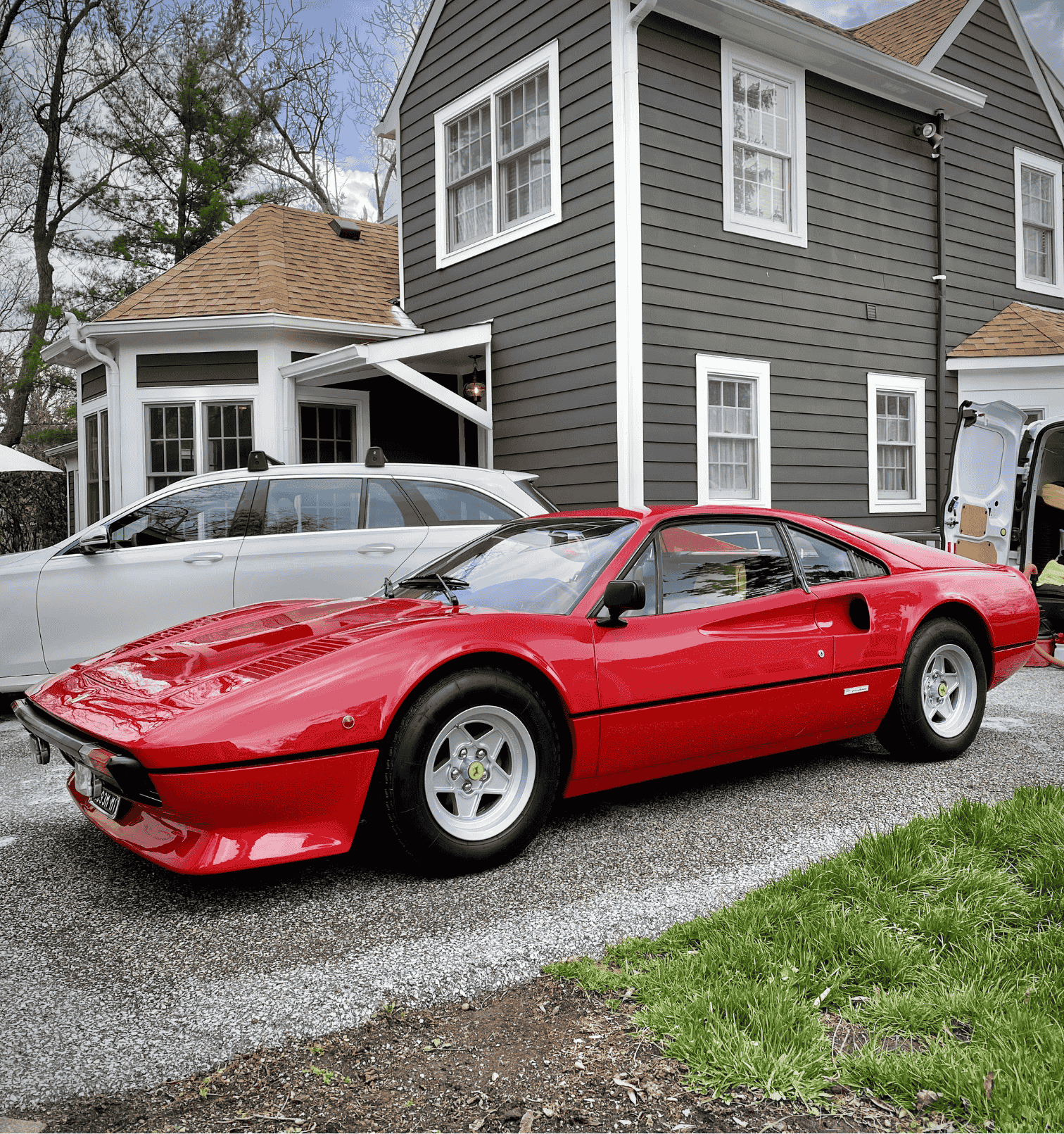 A red vintage sports car parked in a driveway in front of a gray house with white trim and multiple windows, with a silver car and a person unloading items from a white van in the background.