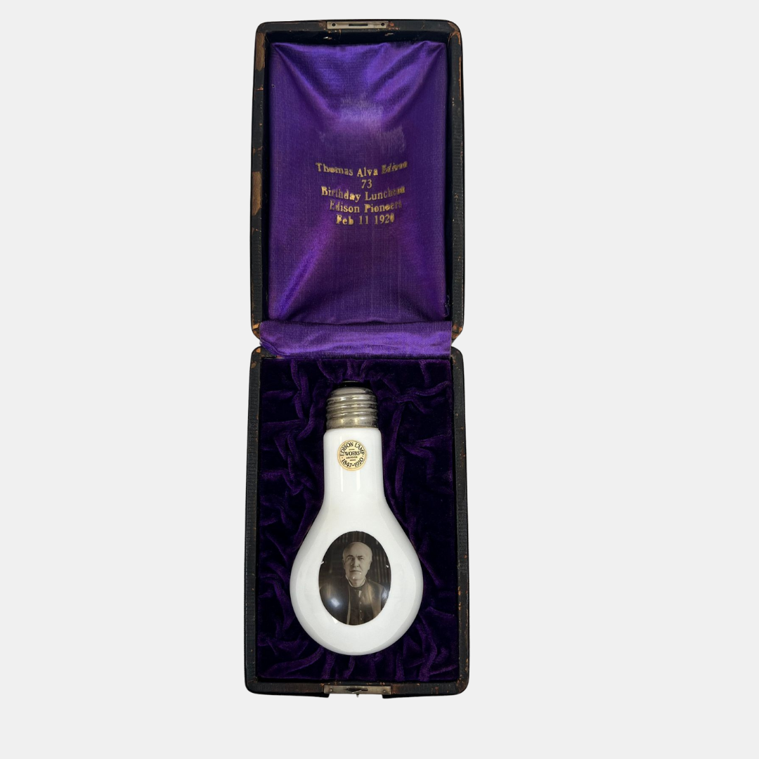 A decorative light bulb with a black and white photo of a man inside it, placed in a black box lined with purple velvet, which has gold lettering inside the lid commemorating a birthday celebration for Thomas Alva Edison on February 11, 1921.
