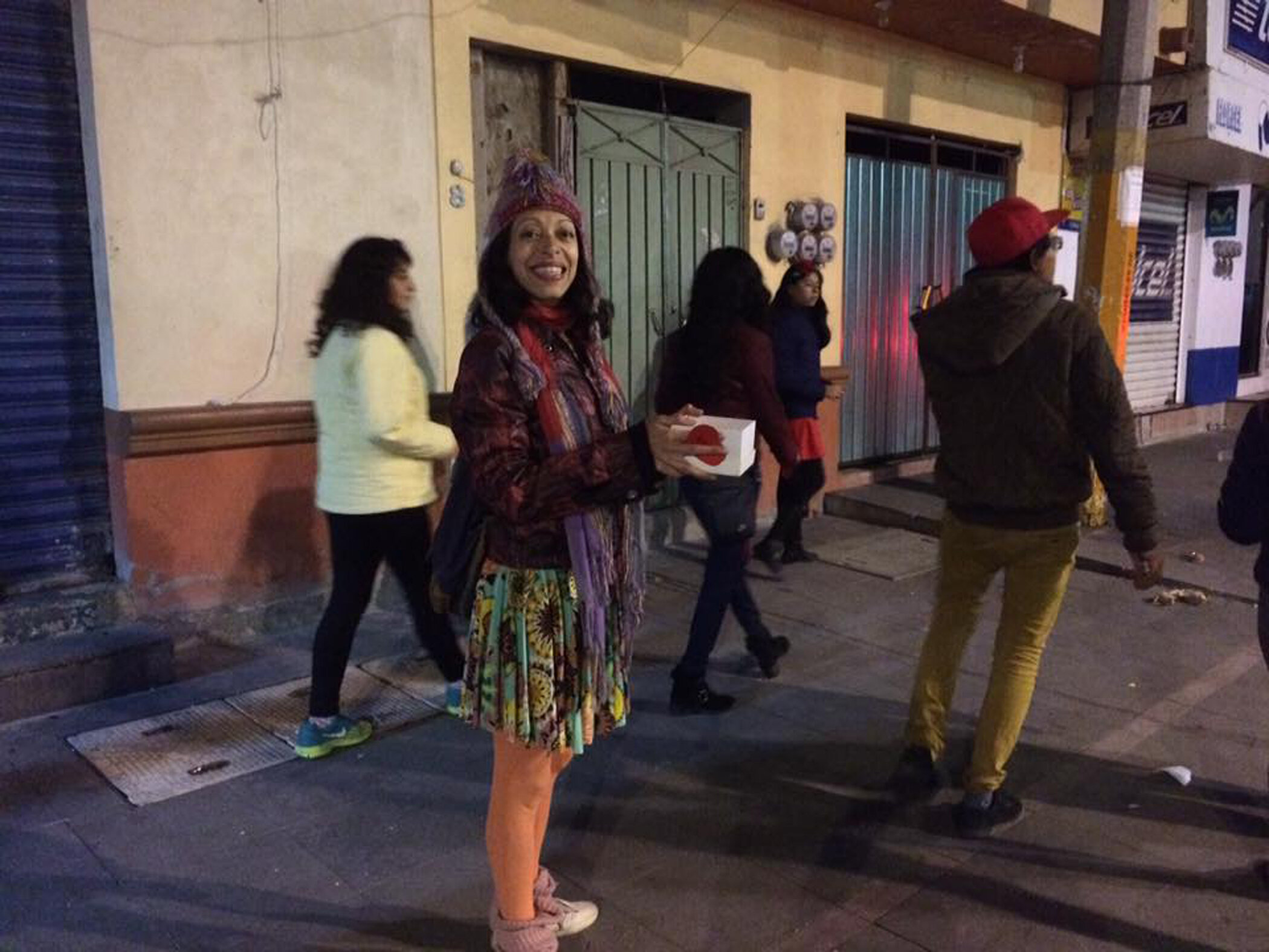 Mujer sonriendo en calle con varias personas caminando al fondo, en un área urbana por la noche.