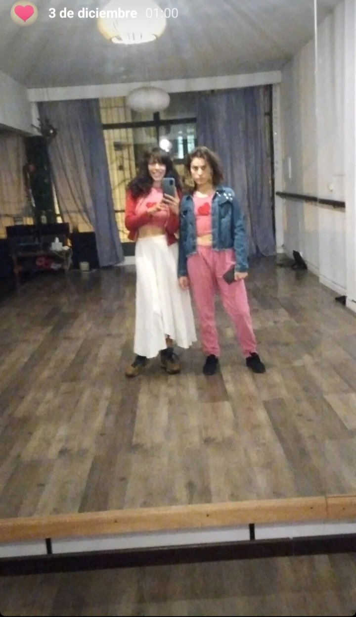Dos mujeres jóvenes tomando una selfie en un espejo grande en una sala con cortinas y lámparas. Una lleva un abrigo rosa y pantalones blancos, la otra una chaqueta de mezclilla y pantalones rosados.