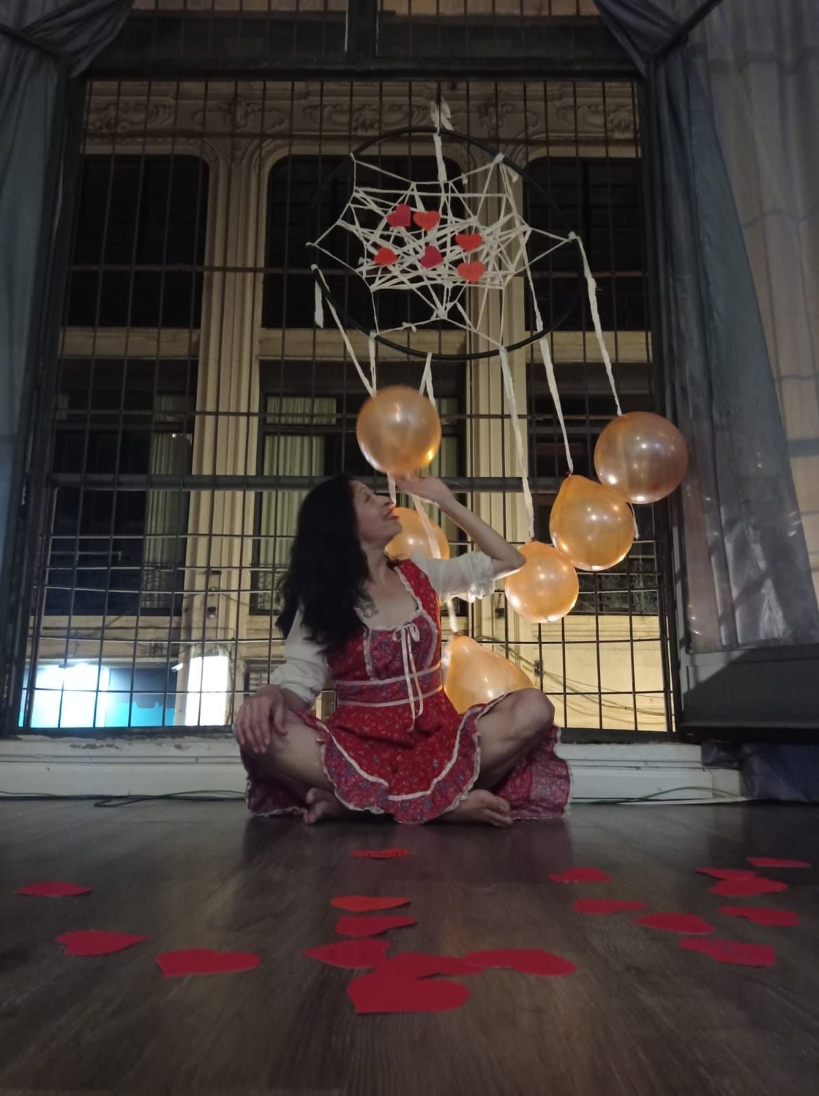 Una mujer sentada en el suelo decorada con globos dorados y corazones rojos, con un ambiente romántico en el fondo.