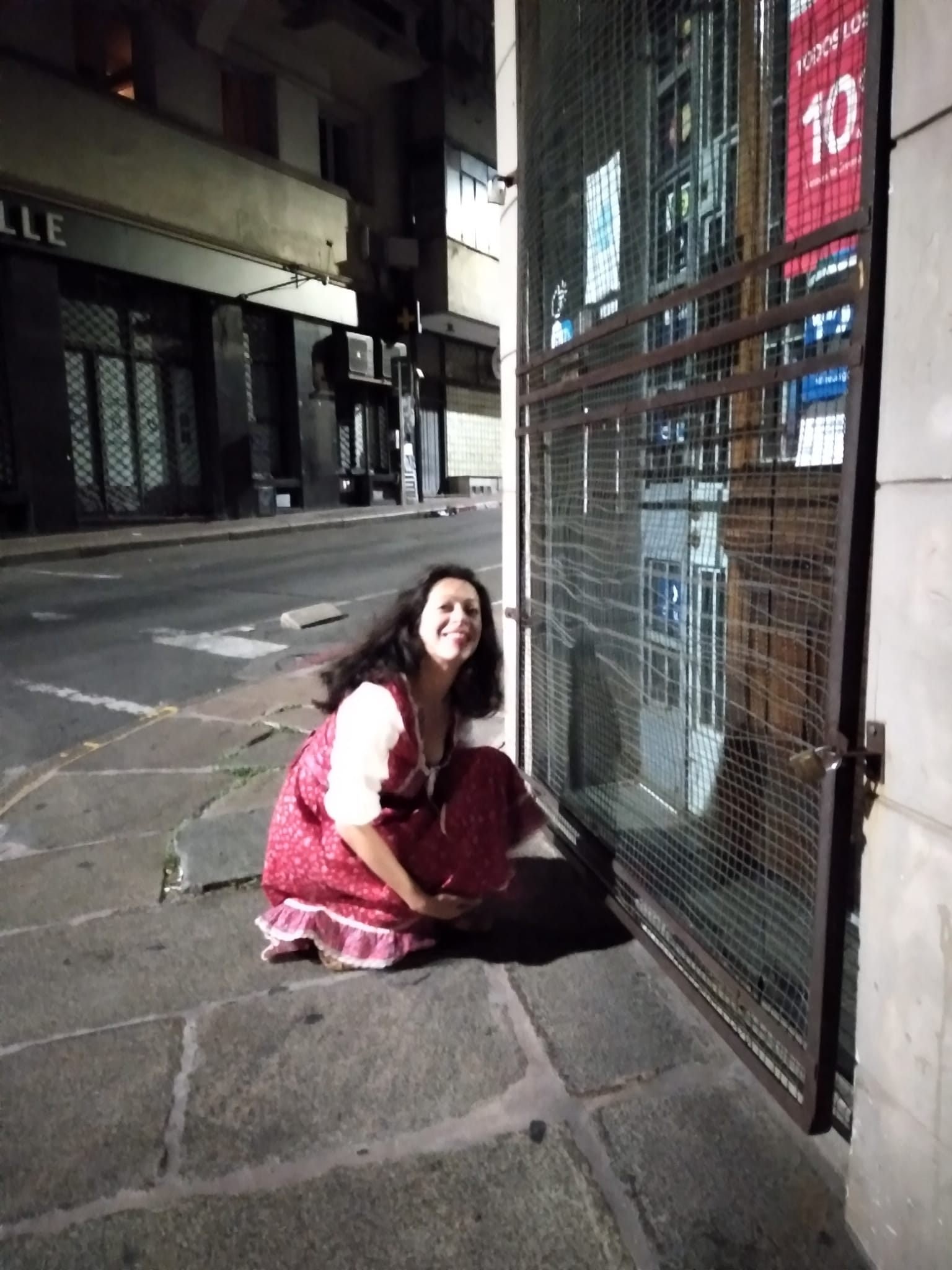 Mujer sonriendo, vestida con falda y blusa blanca, agachada junto a una puerta de reja en la calle por la noche.