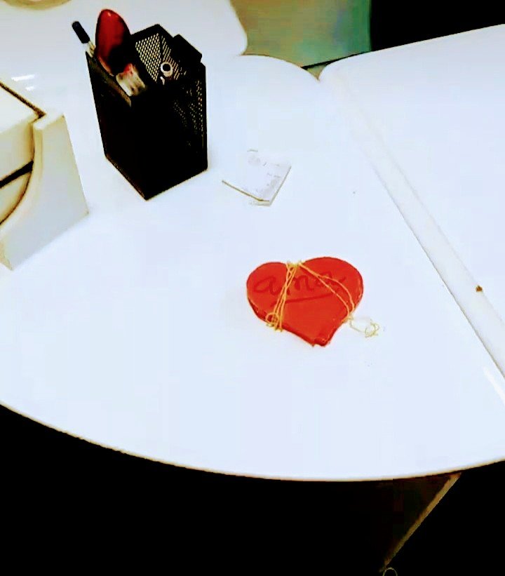 Corazón de papel rojo con la palabra 'Amor' escrito en él, atado con hilo dorado, sobre una mesa blanca, con un organizador de papel negro con archivos y una libreta.