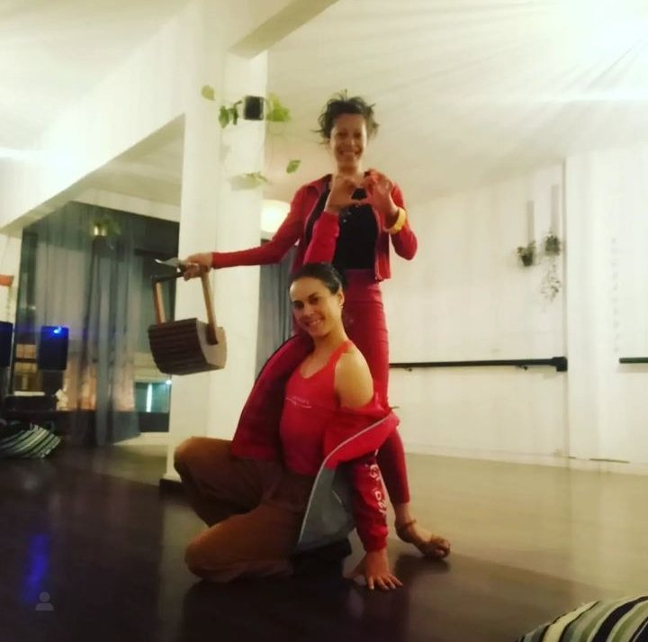 Dos mujeres jóvenes posando en un espacio interior con madera en el suelo, una de ellas sentada y la otra de pie, ambas sonriendo y vestidas con ropa roja y negra.
