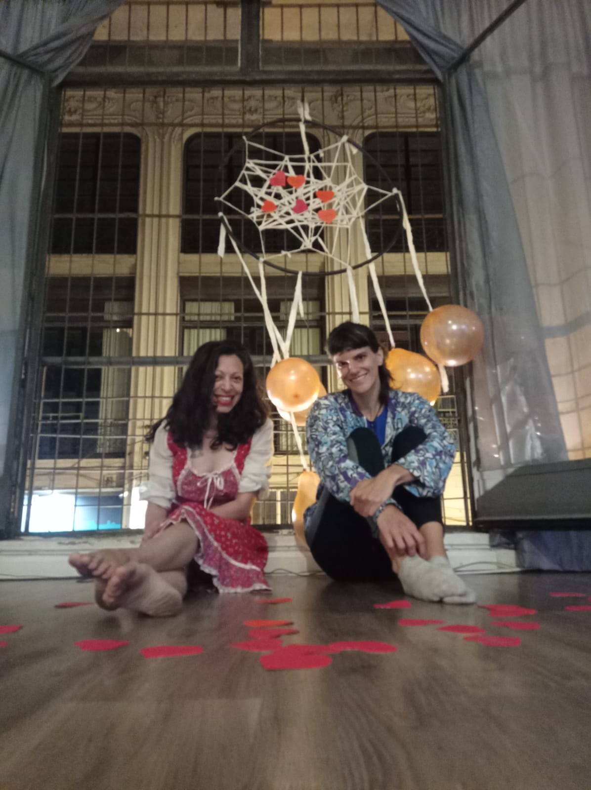Dos personas sentadas en el suelo en una festividad, decoradas con confeti rojo y una estructura decorativa con globos, telaraña y corazones en el fondo en un espacio de interior.