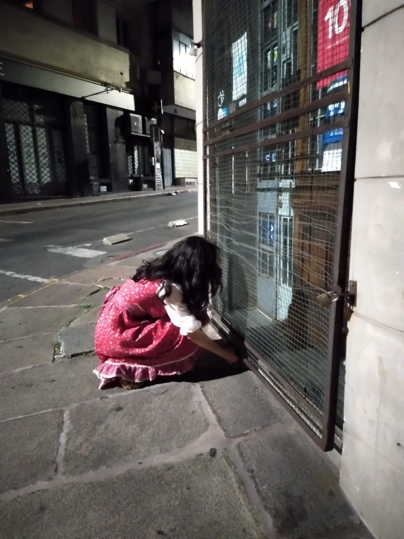 Niña con vestido rosa, de espaldas, enojando una reja en la acera de la calle por la noche.