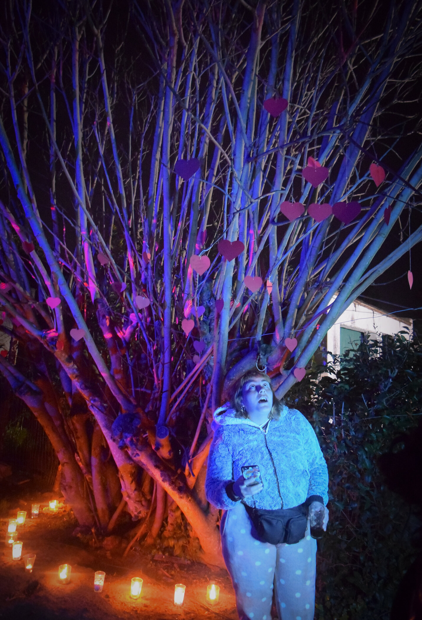 Mujer con pijama de lunares y teléfono móvil en mano, frente a un árbol con corazones de papel, en una noche iluminada con linternas y luces de colores