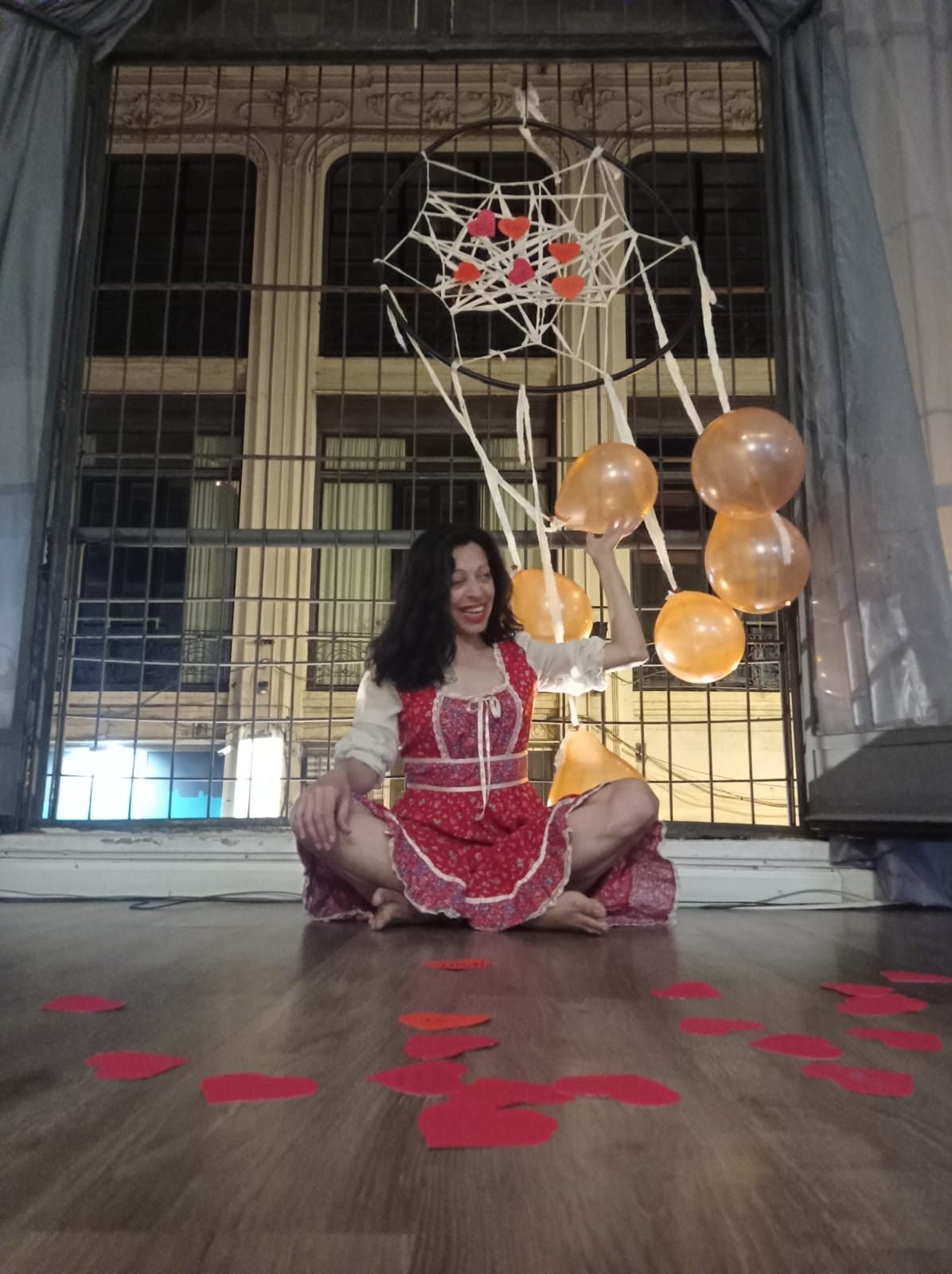 Mujer con vestido rojo sentado en el piso con decoraciones rojas, doradas y de telaraña en el fondo, sonriendo.