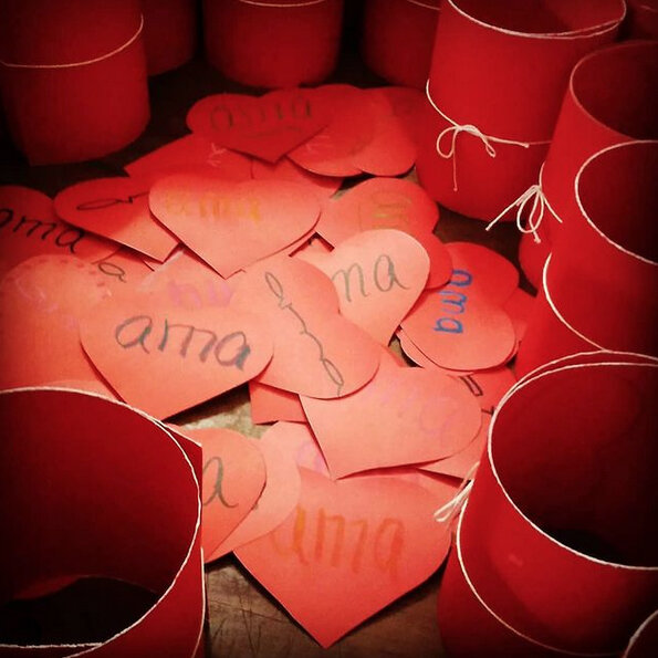 Corazones de papel hechos a mano con diferentes palabras y frases, rodeados por cilindros rojos de cartulina o papel, en un ambiente de manualidades o decoración.