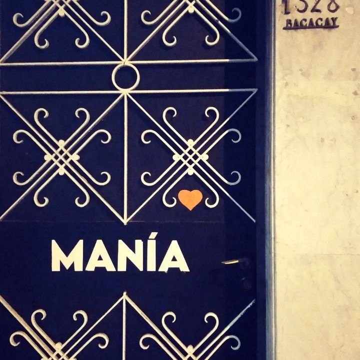 Puerta azul con decoraciones blancas y la palabra 'MANÍA' en letras grandes. Hay un corazón naranja sobre la letra I.