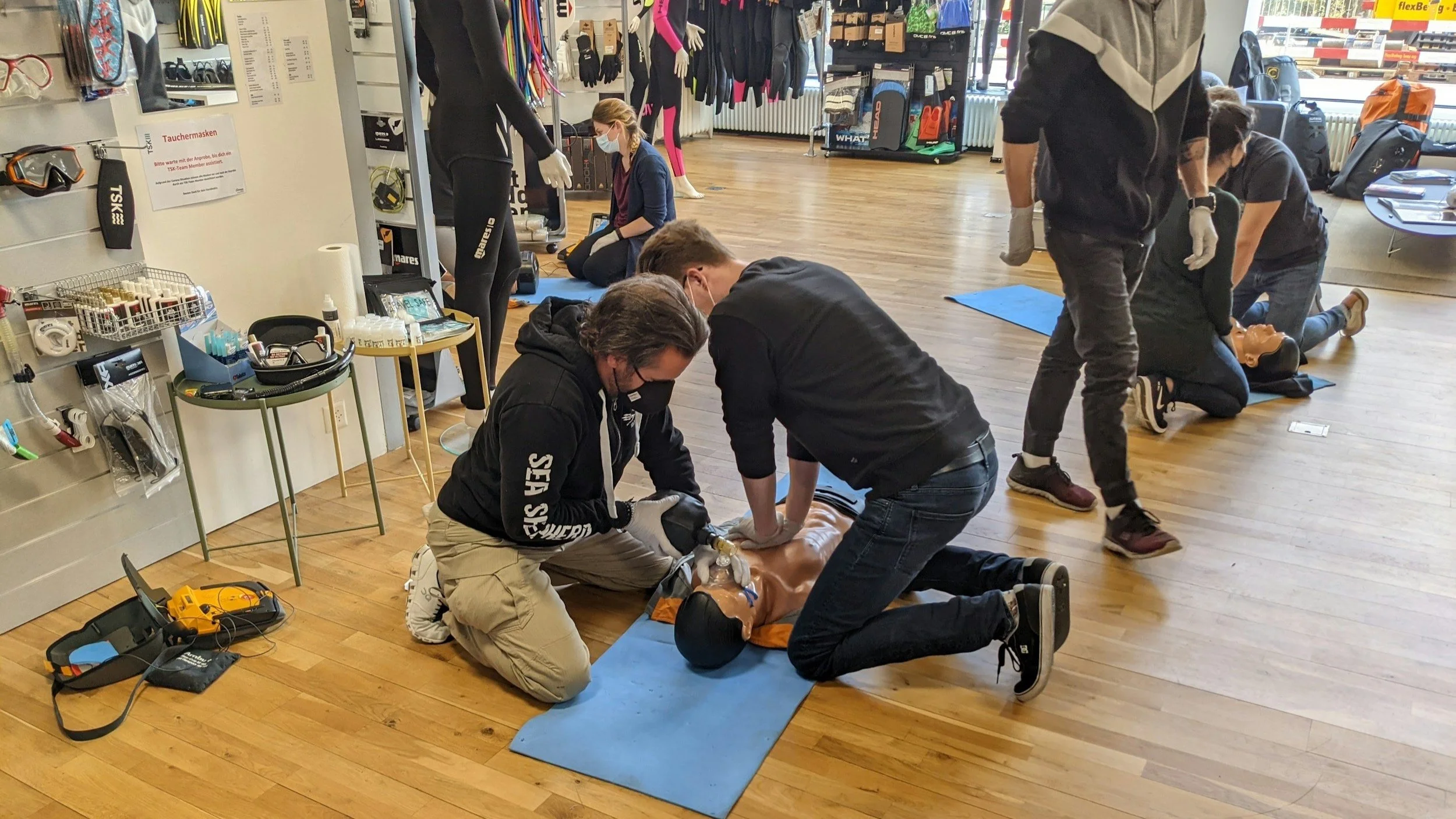 A CPR training session with multiple people practicing on mannequins inside a store or fitness shop. Two men are performing CPR on a mannequin on the floor, with other participants practicing nearby.