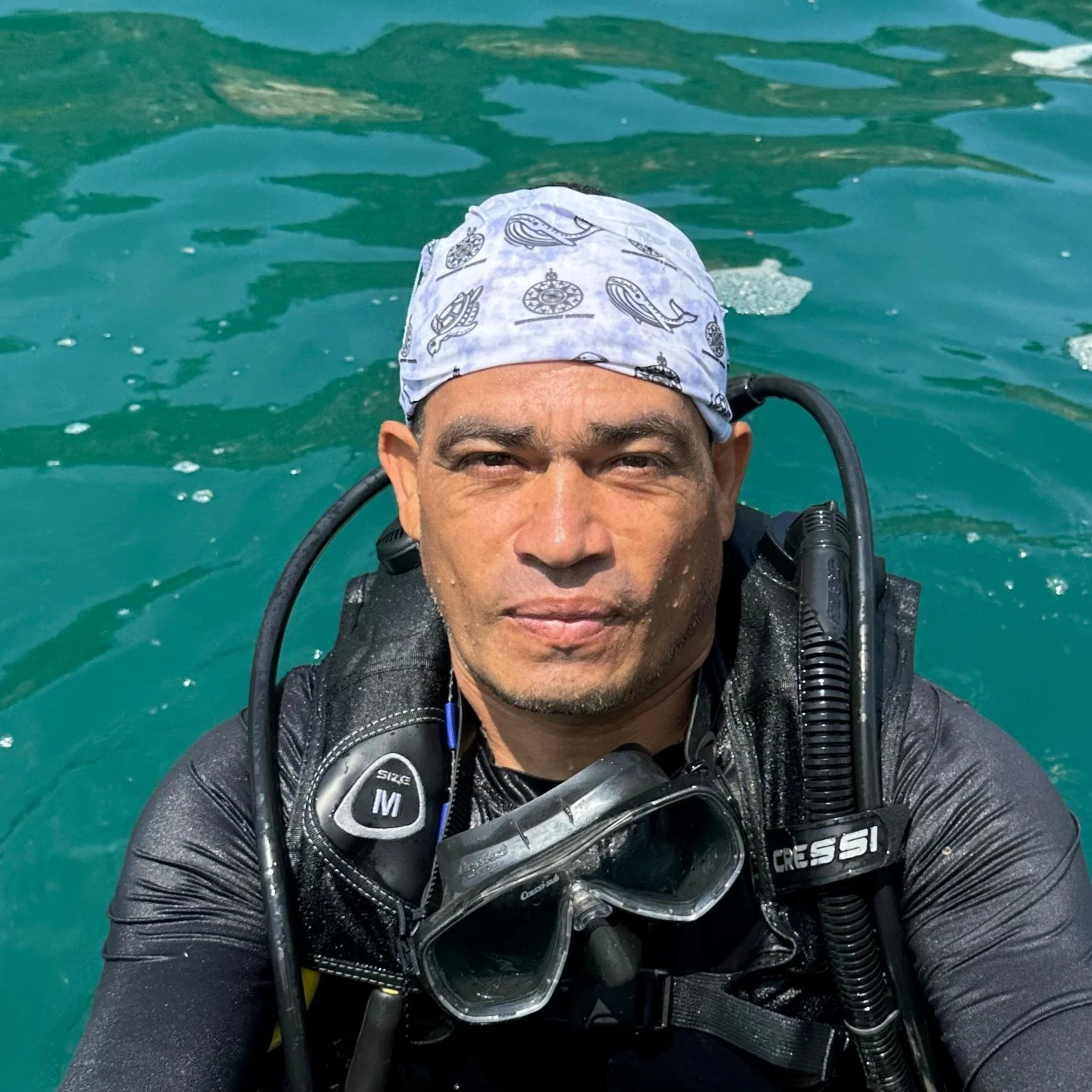 A man in scuba gear, with a white bandana, taking a selfie in the water.