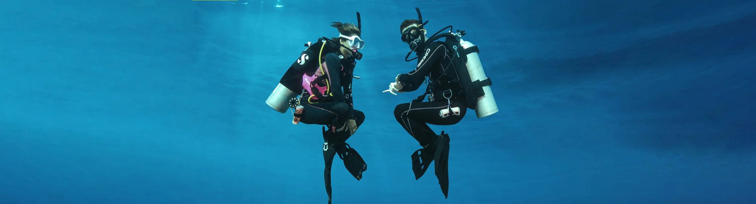 Two scuba divers underwater, facing each other, wearing diving suits and equipment including tanks and masks.