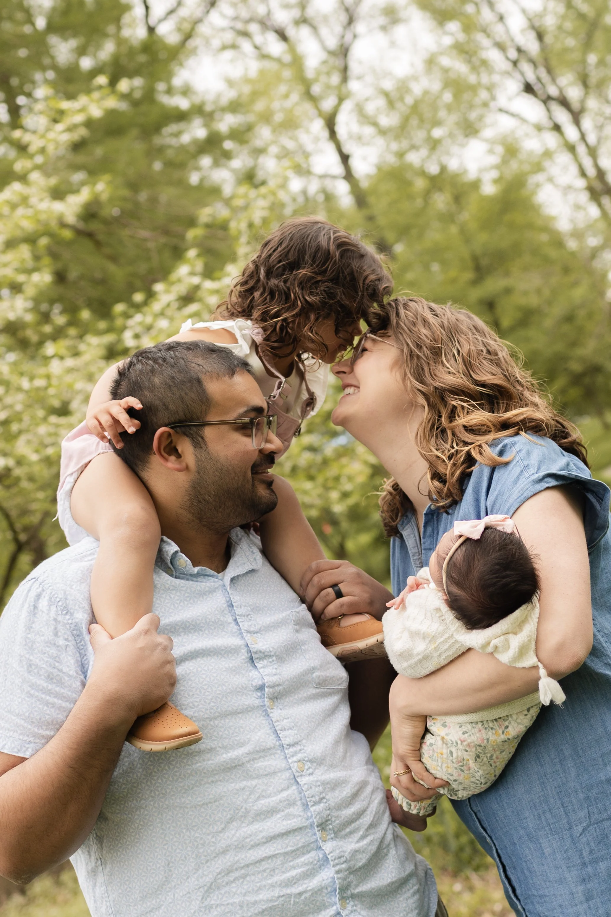 A family of four outdoors, with a man, woman, young girl, and baby, touching foreheads and smiling, in a park with green trees.