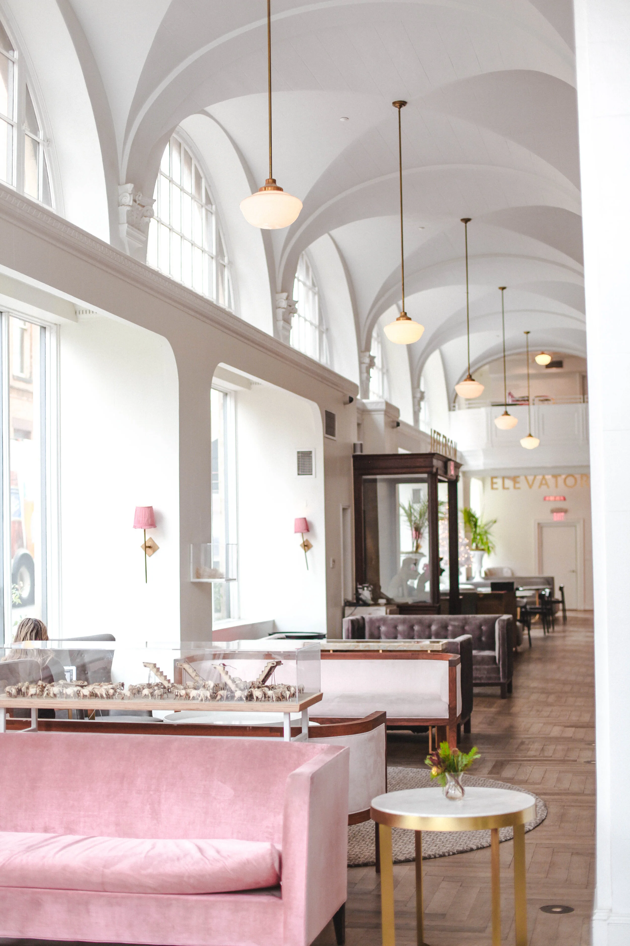 Interior of a bright, spacious lounge or lobby with white arched ceilings, large windows, pink velvet sofa, small round table with a vase of flowers, and pendant lights hanging from ceiling.