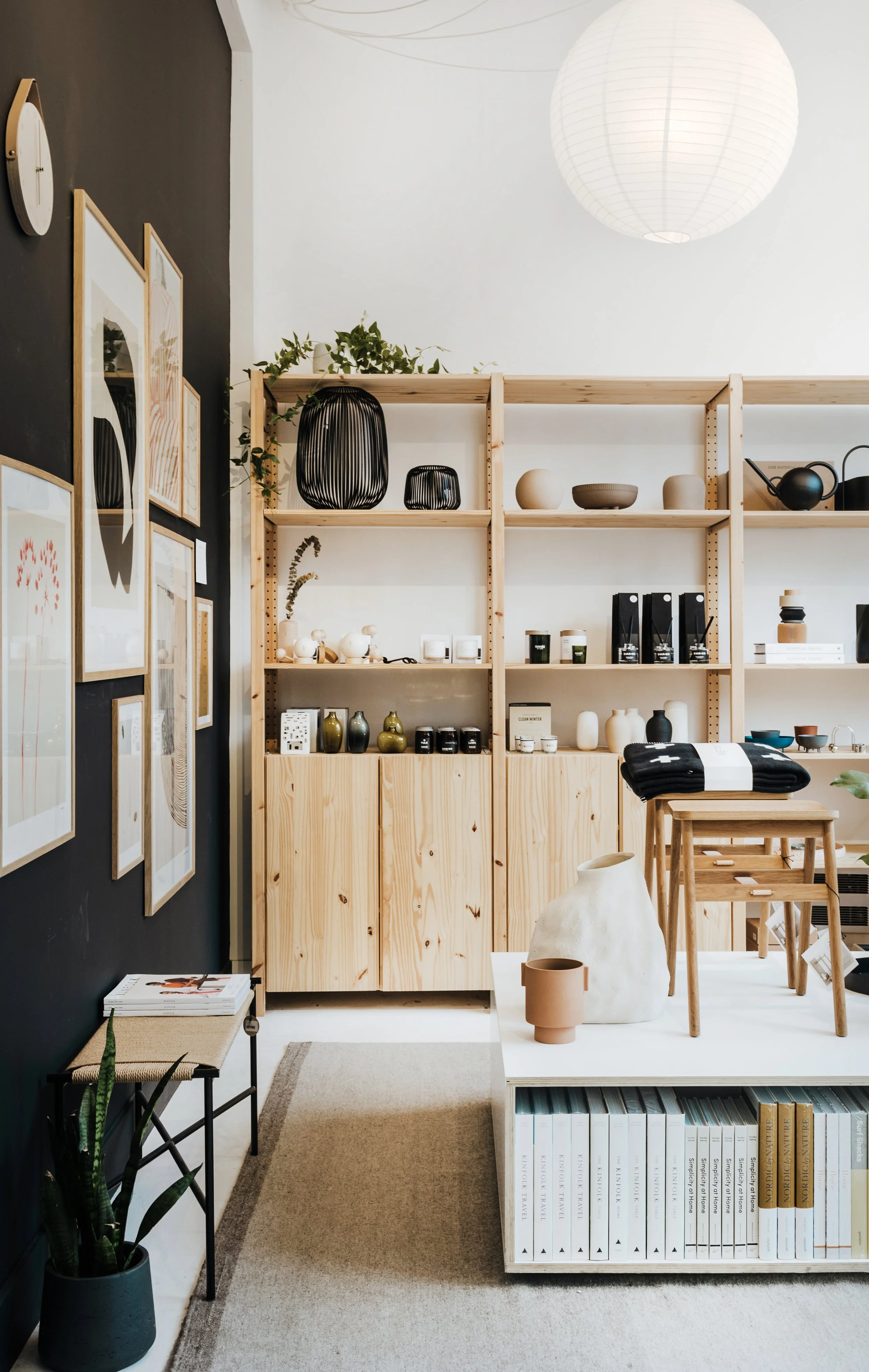 Interior of a modern home decor store with wooden shelves holding pottery, candles, and decorative objects, a black accent wall with framed art, a small bench, a potted plant, and a white display table with more vases and books.