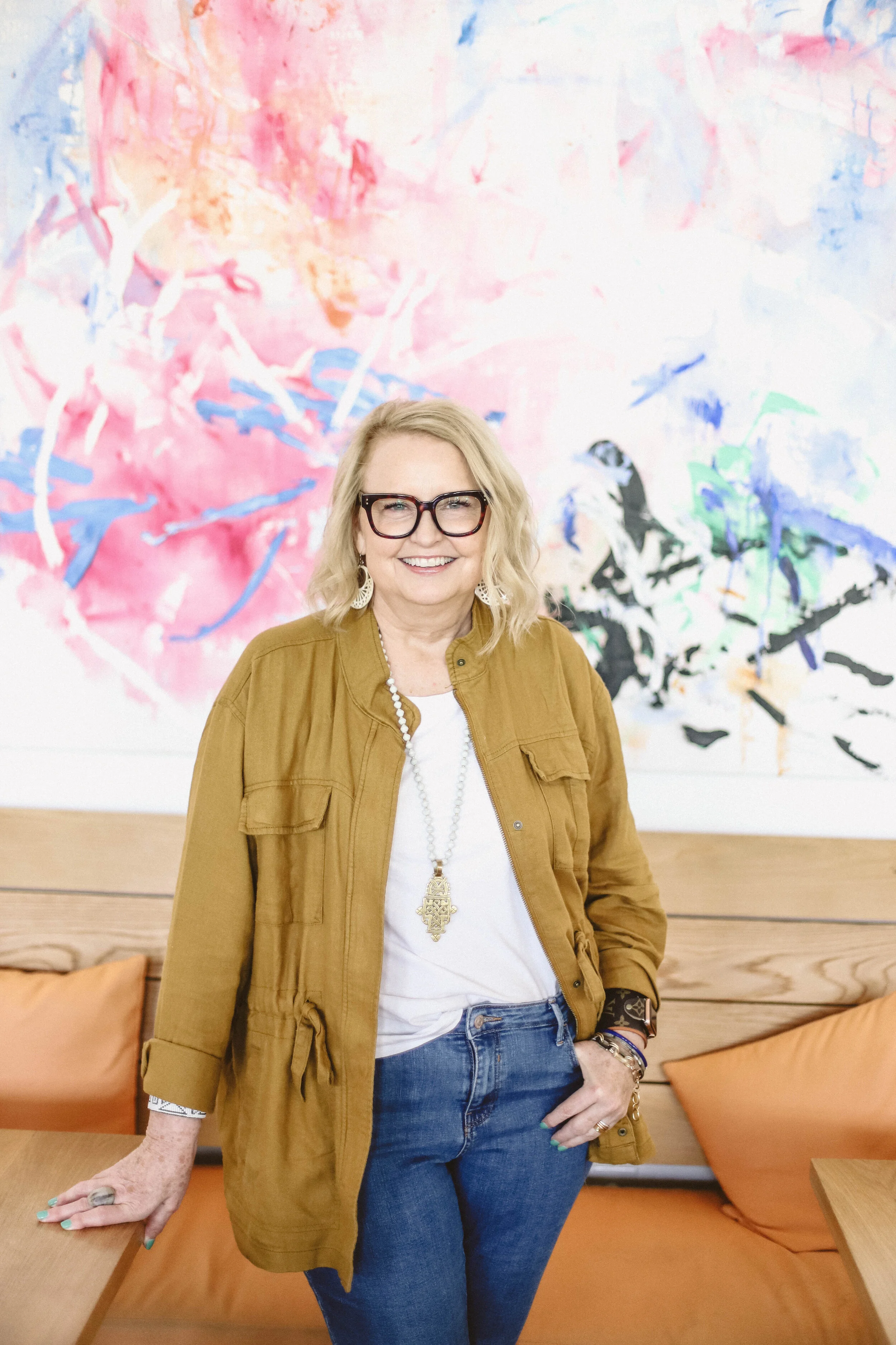 A woman with blonde hair, glasses, and light skin is smiling and standing in front of a colorful abstract painting. She is wearing a mustard jacket, white top, blue jeans, and jewelry.