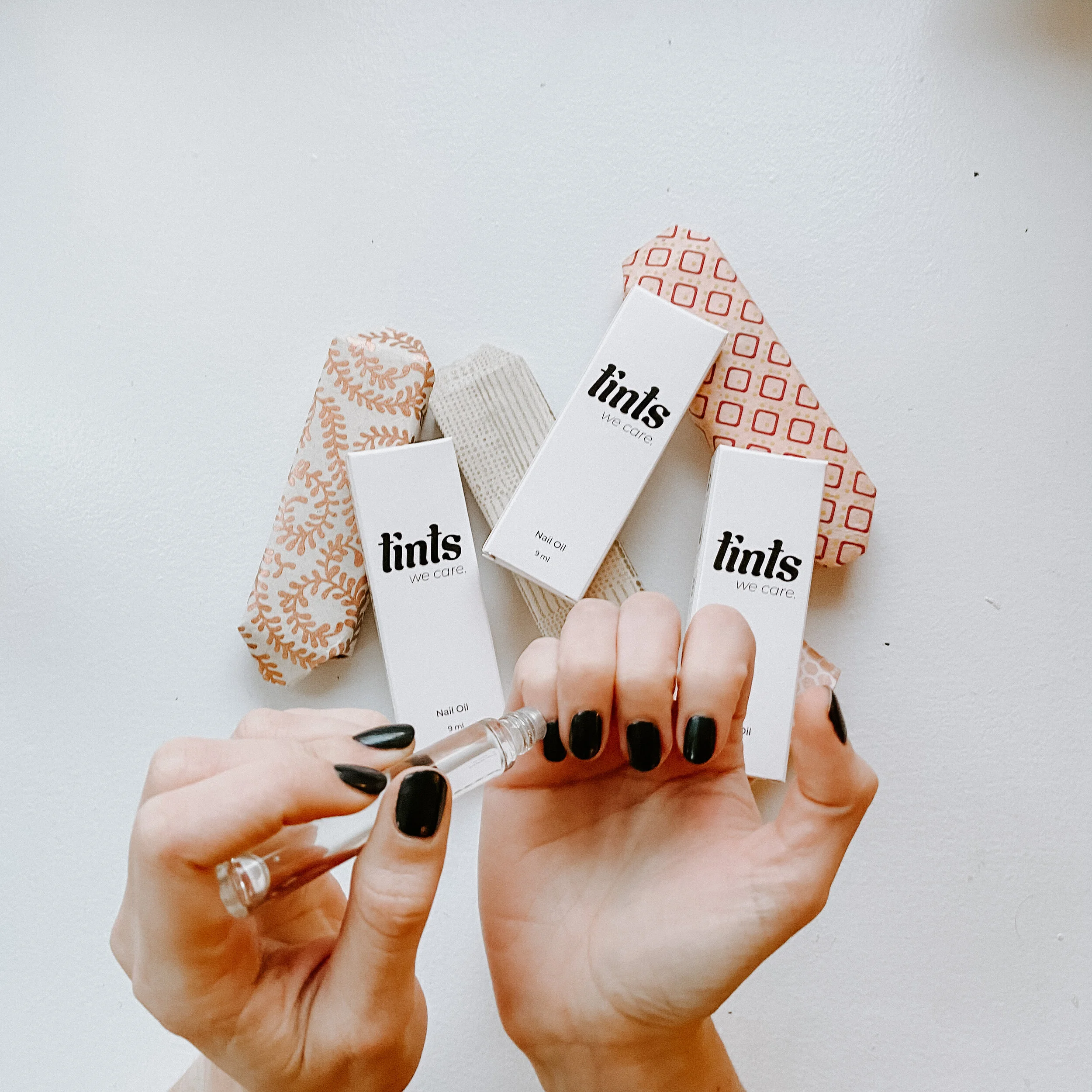 Hands with black nail polish holding a small bottle of nail oil over a white surface, surrounded by three boxes of Tints nail care products and patterned wraps.