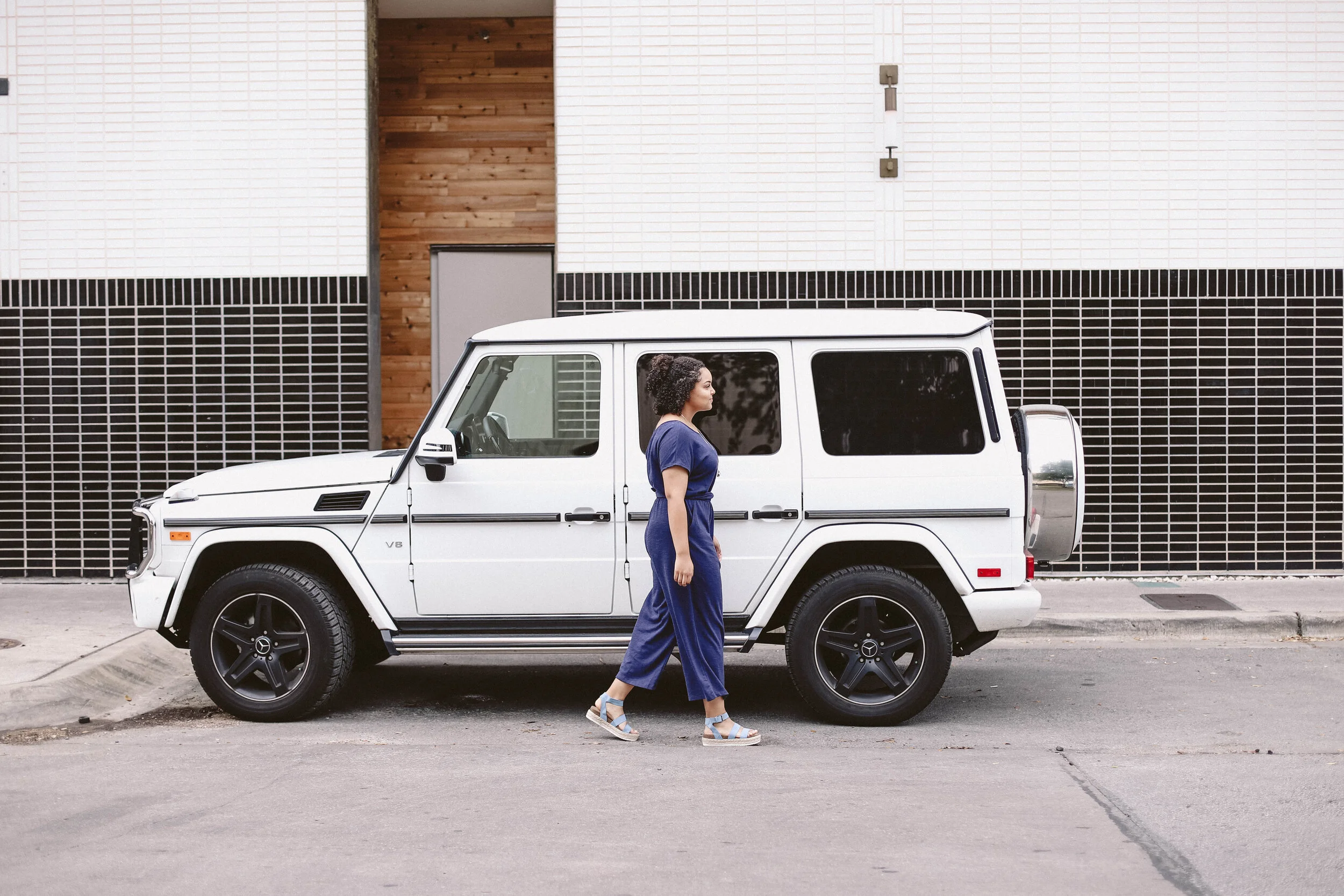 A woman with curly hair dressed in a blue jumpsuit and slip-on sandals walking alongside a white Mercedes-Benz G-Class SUV parked on a city street.