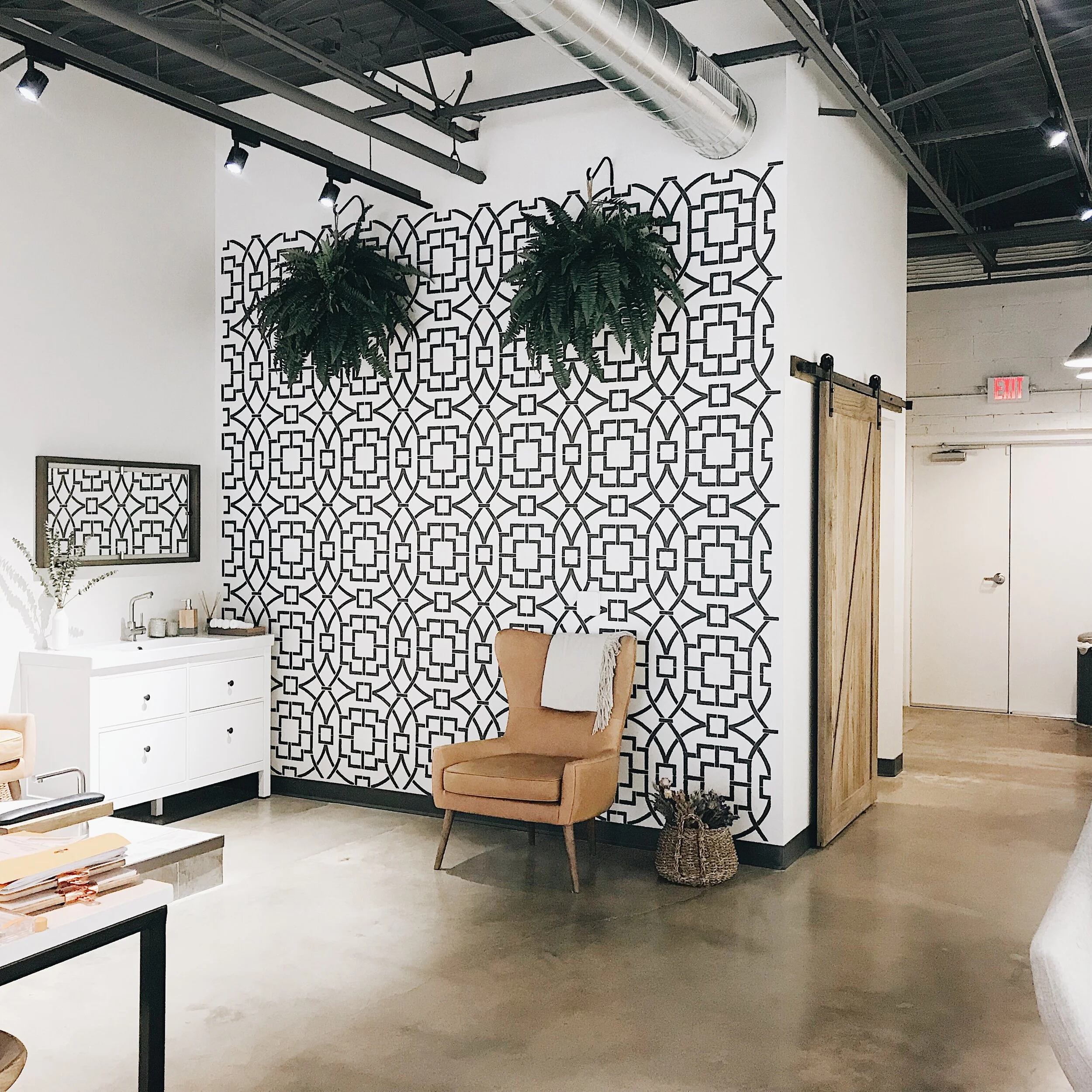 Interior space with a wall decorated with a black and white geometric pattern, a beige armchair with a white blanket, a white cabinet with a mirror above it, and hanging green plants, industrial ceiling with ductwork.