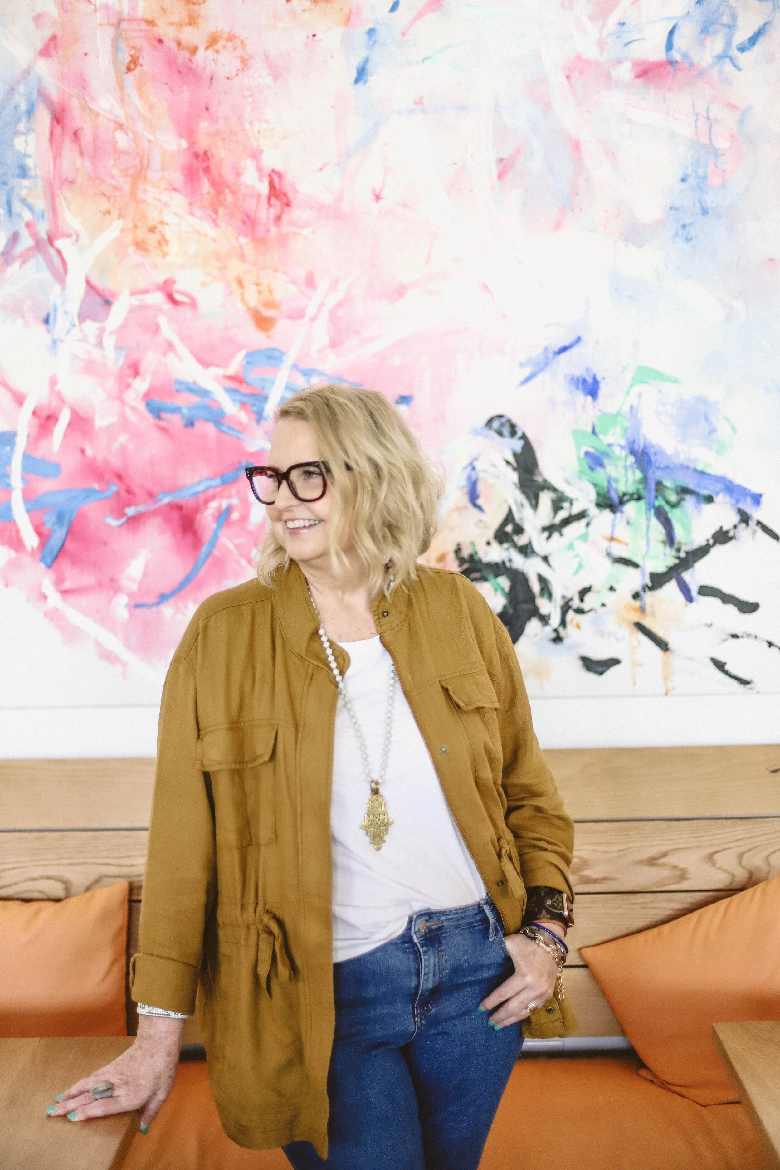 Woman with blonde curly hair, glasses, white shirt, mustard coat, and jeans, smiling and standing in a room with colorful abstract art on the wall behind her and orange cushions on a wooden bench.