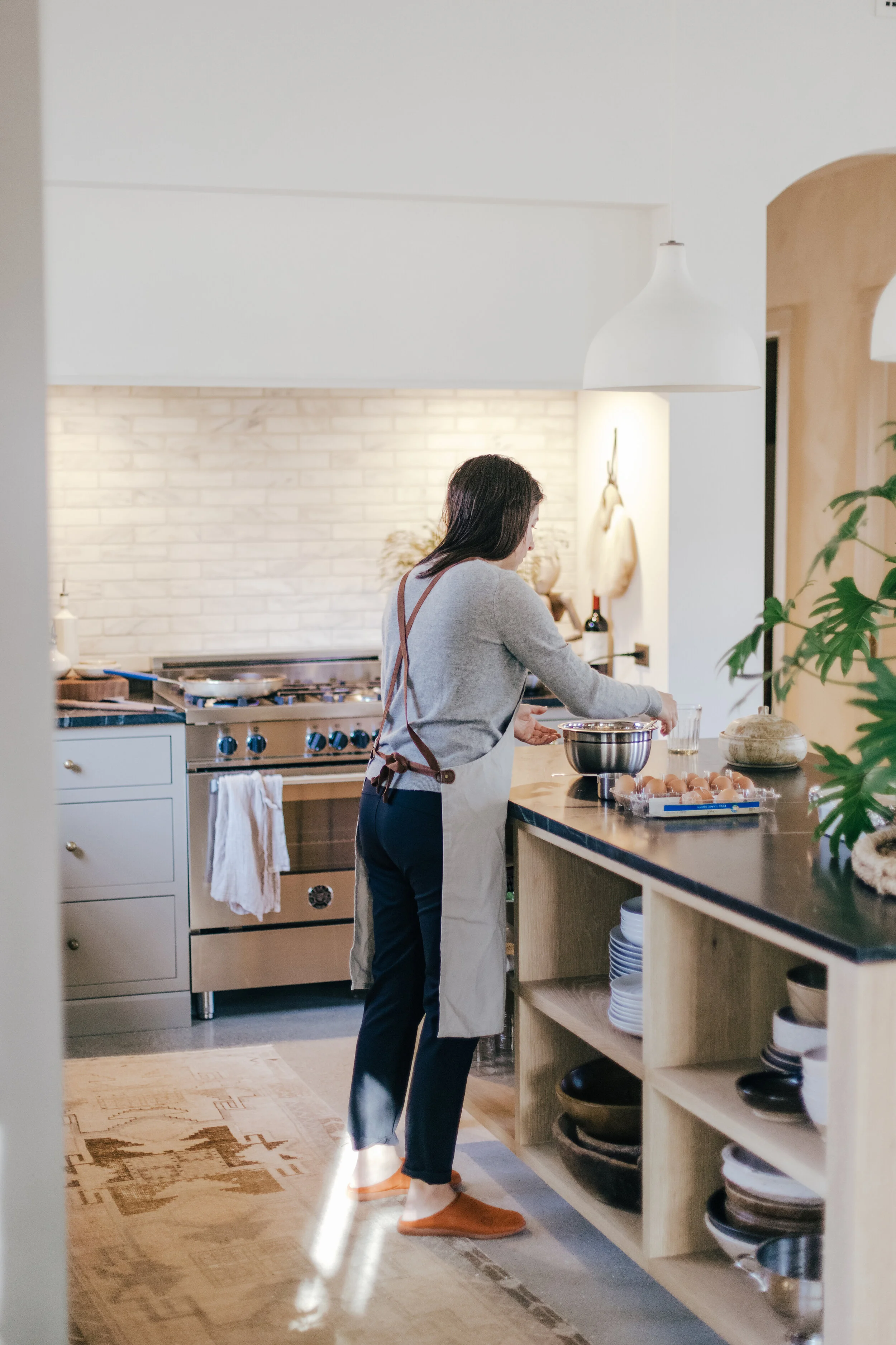 Woman in a gray sweater and navy pants, wearing an apron, preparing food in a modern kitchen with open shelves, a stainless steel stove, and decorative items.