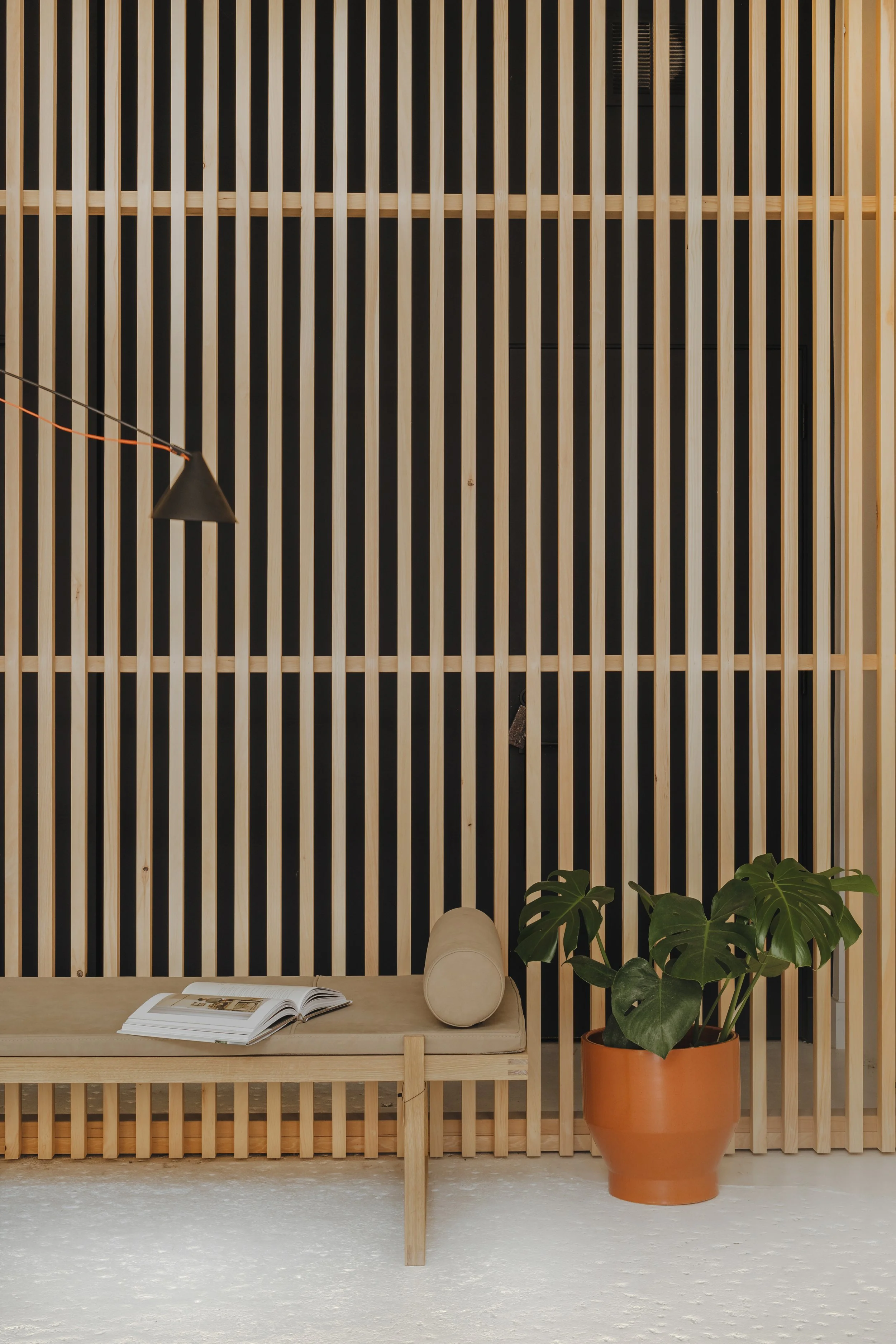 Interior space featuring a wooden slatted wall, a beige bench with an open book and cylindrical pillow, a large potted Monstera plant, and a black wall-mounted lamp.
