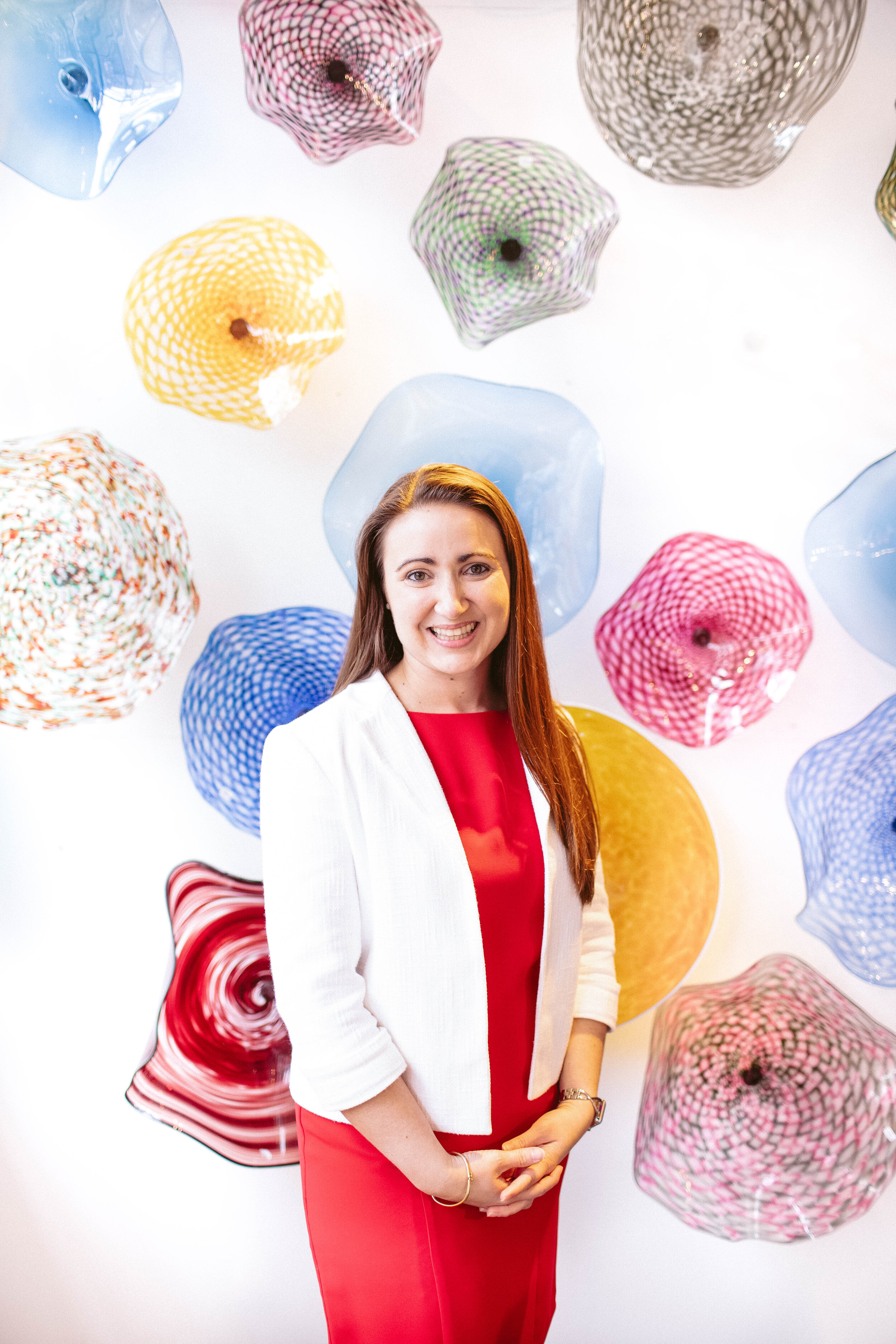 A woman with long brown hair wearing a red dress and white blazer standing in front of a colorful abstract art installation of glass sculptures hanging from the ceiling.