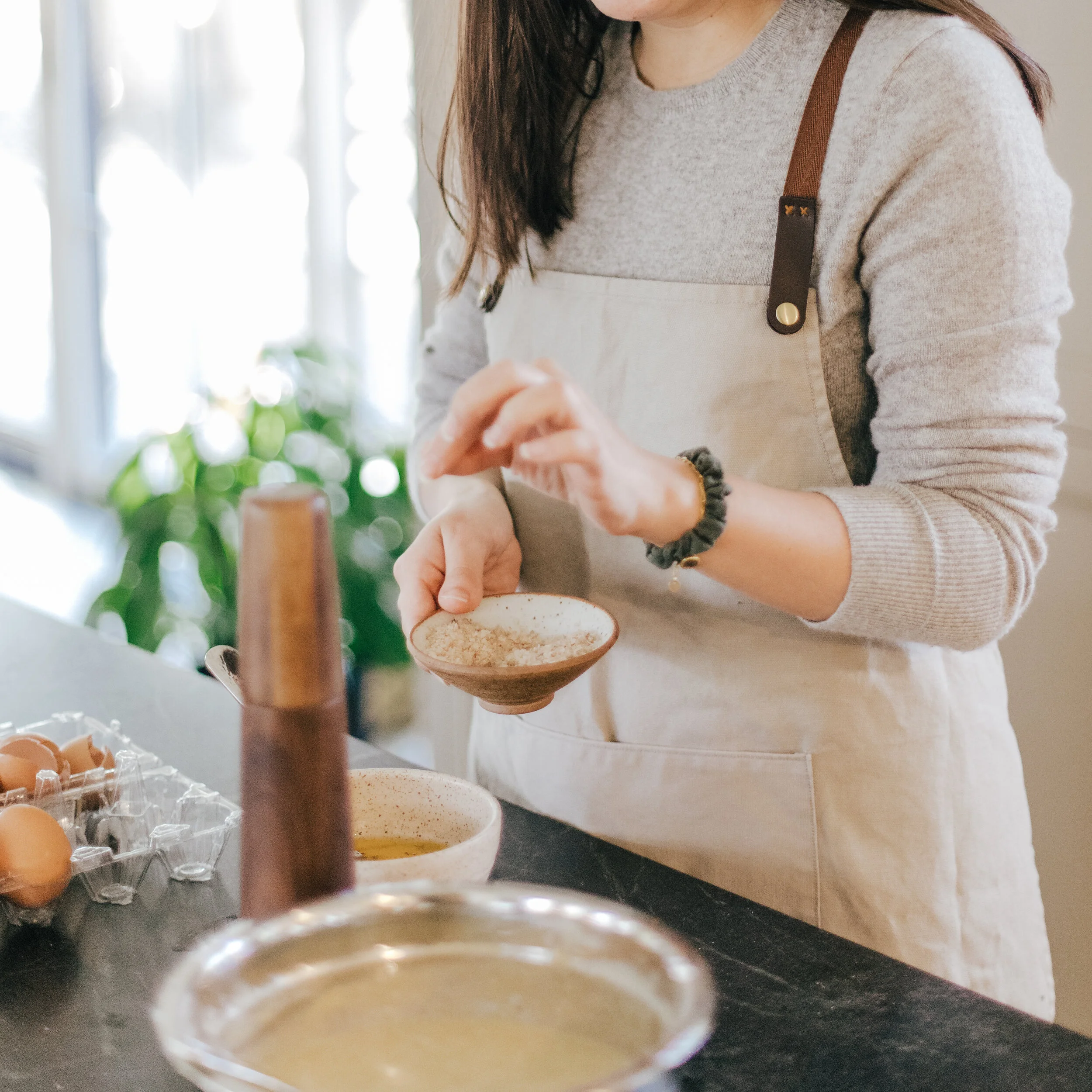 A woman wearing a beige apron and gray sweater is cooking in a kitchen, pouring ingredients into a small bowl. There are eggs and mixing bowls on the countertop.