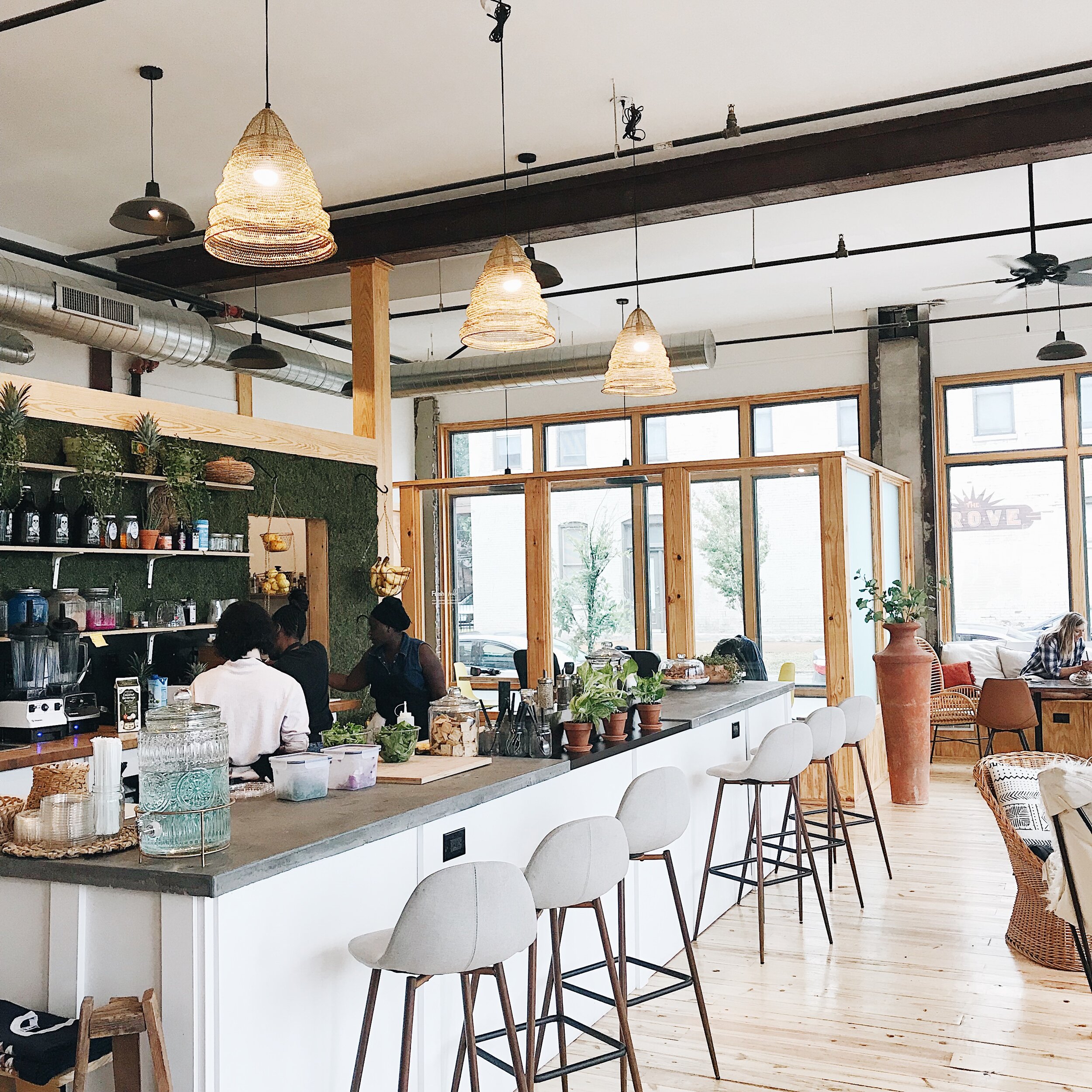 Interior of a modern cafe with a bar counter, white chairs, potted plants, large windows, and hanging woven light fixtures.