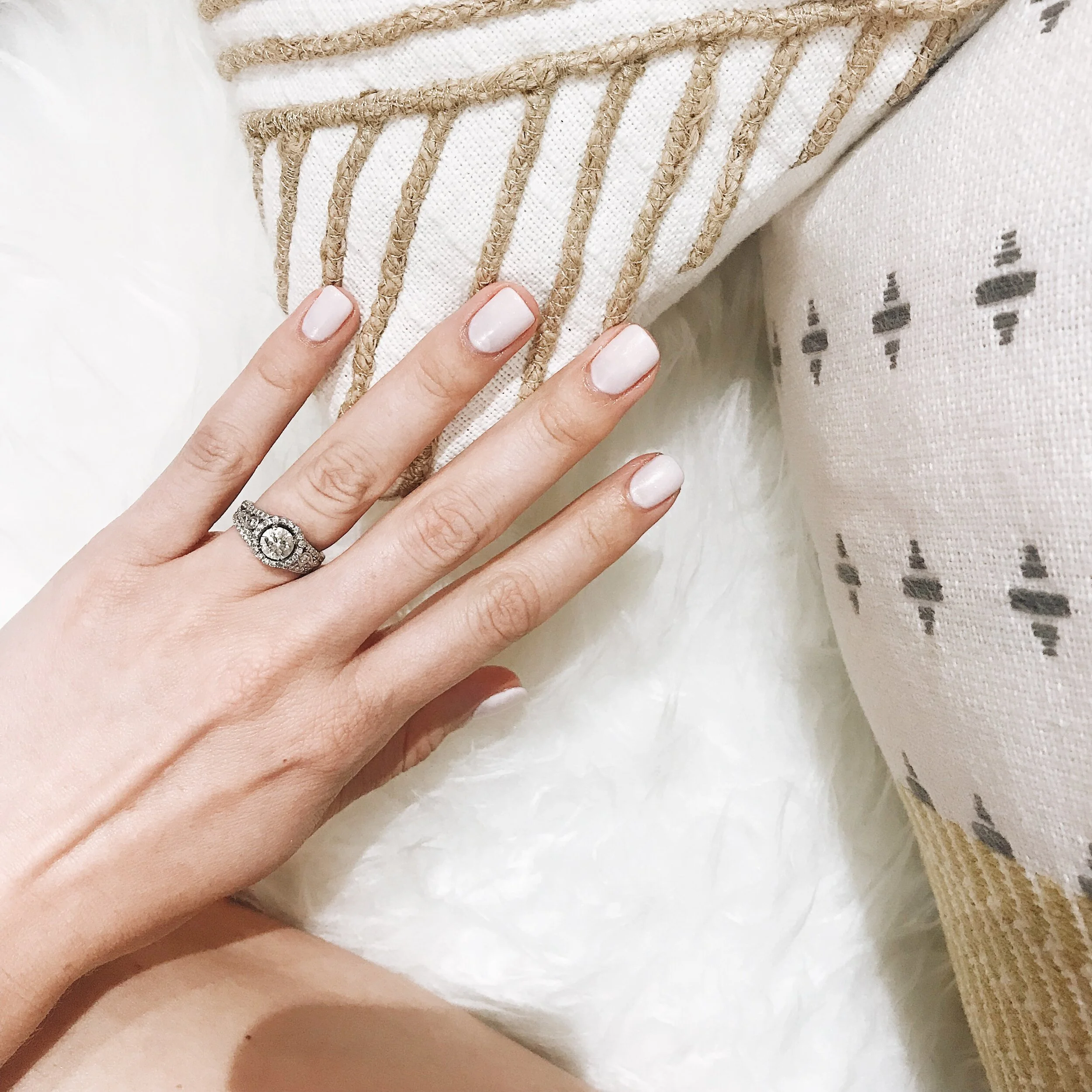 Close-up of a hand with a diamond ring resting on decorative pillows with striped and embroidered patterns on a white fluffy surface.