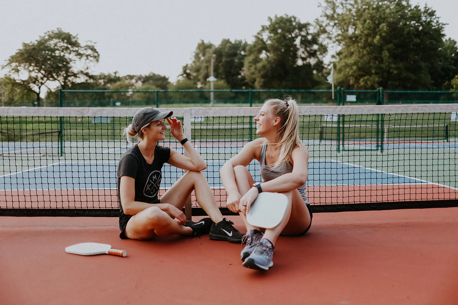 Two women sitting on a tennis court near the net, having a conversation and smiling, with Recess Pickleball paddles in Tower Grove Park on the ground beside them in St. Louis, MO.