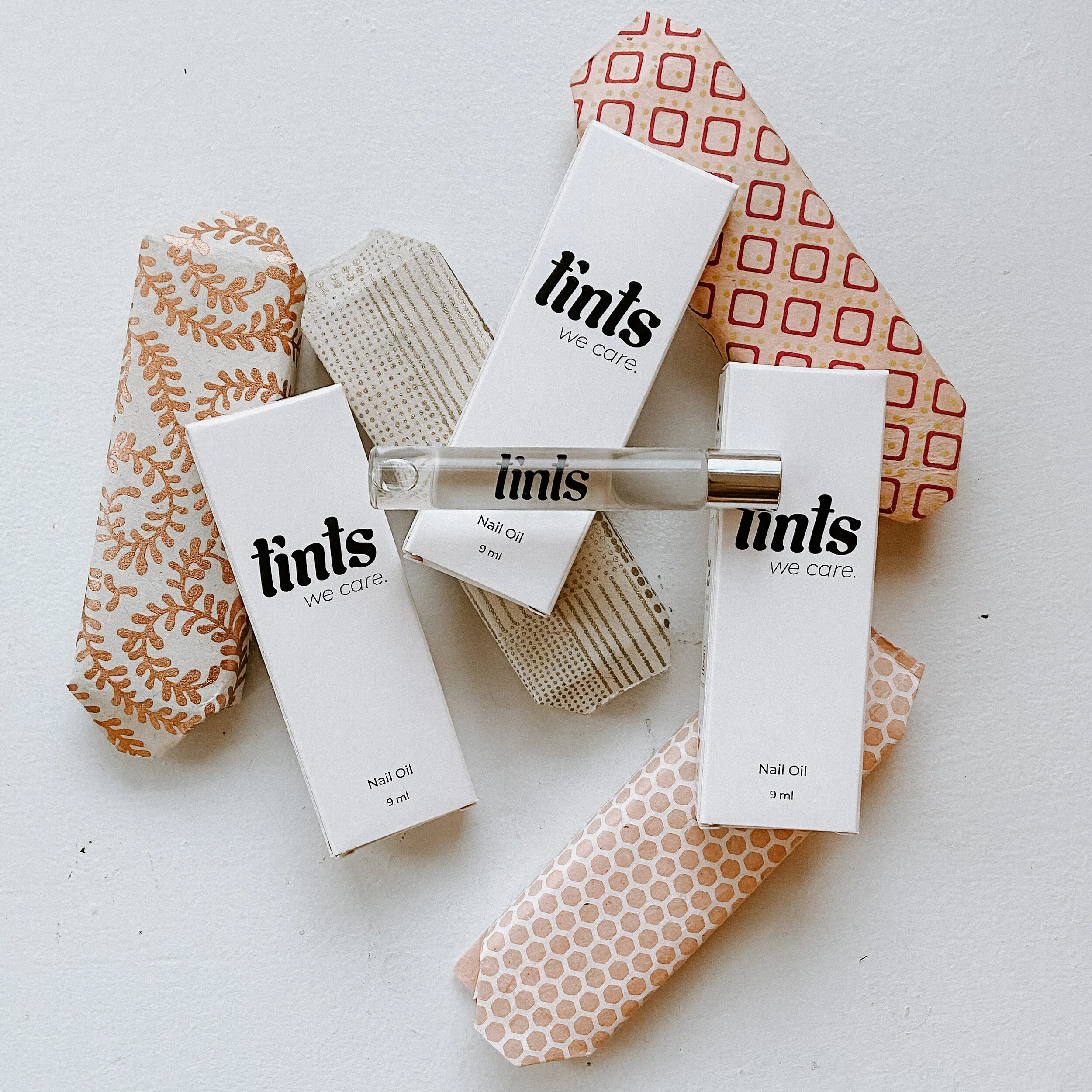 Multiple boxes of Tints nail oil with a small glass roller bottle on paper towels decorated with various geometric and leaf patterns.