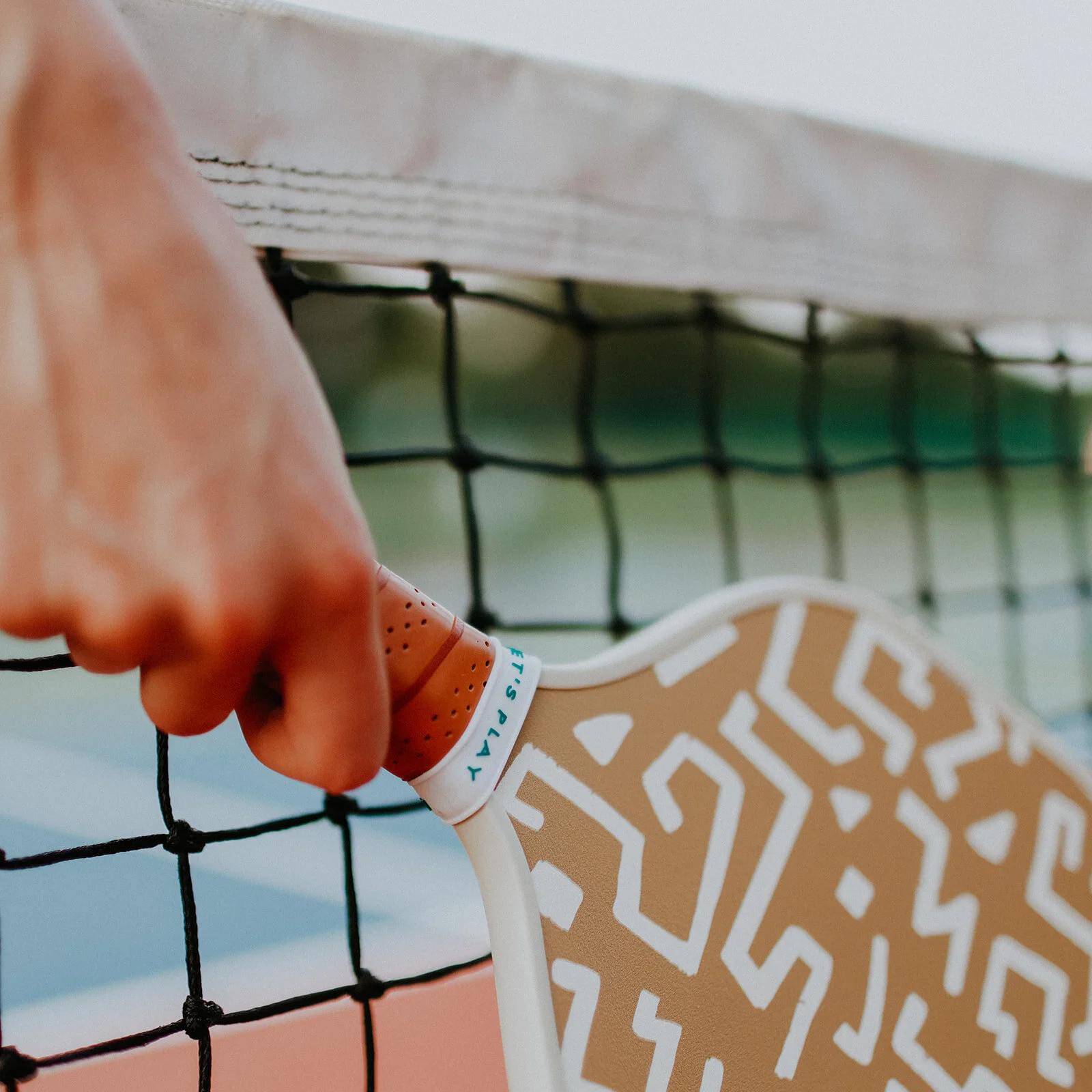 Close-up of a person's hand holding a Recess Pickleball paddle in Tower Grove Park at the net on a tennis court.