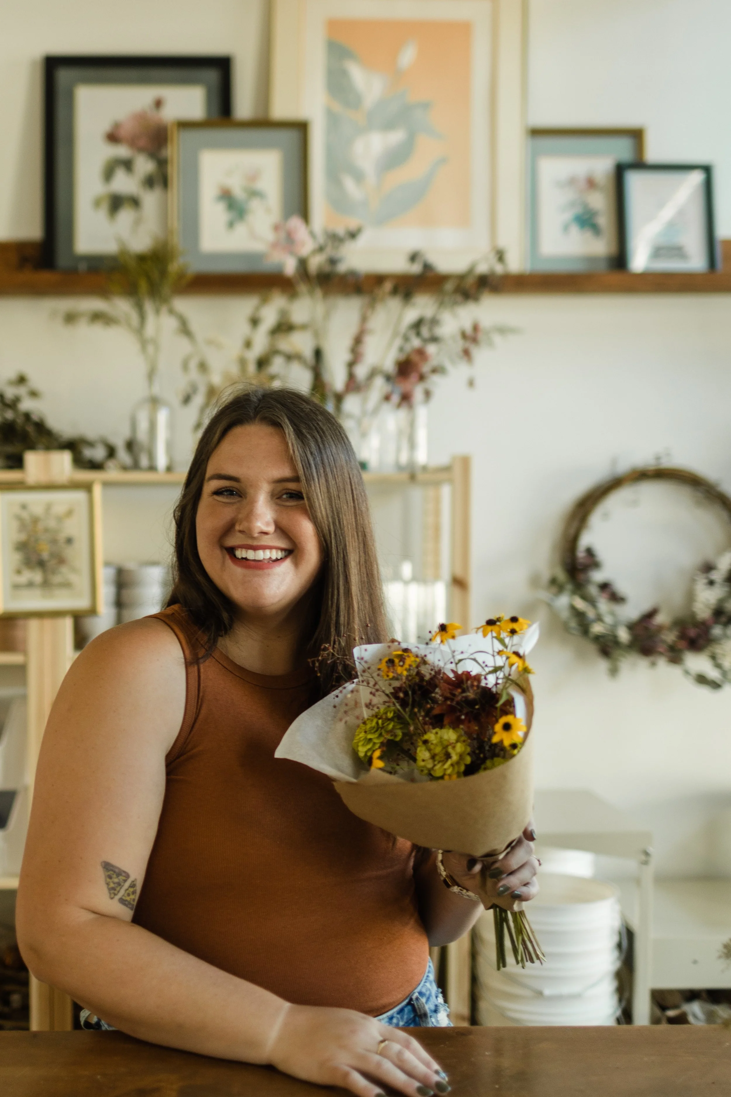 A smiling woman holding a bouquet of yellow and red flowers inside a flower shop decorated with framed artwork, floral arrangements, and wreaths.